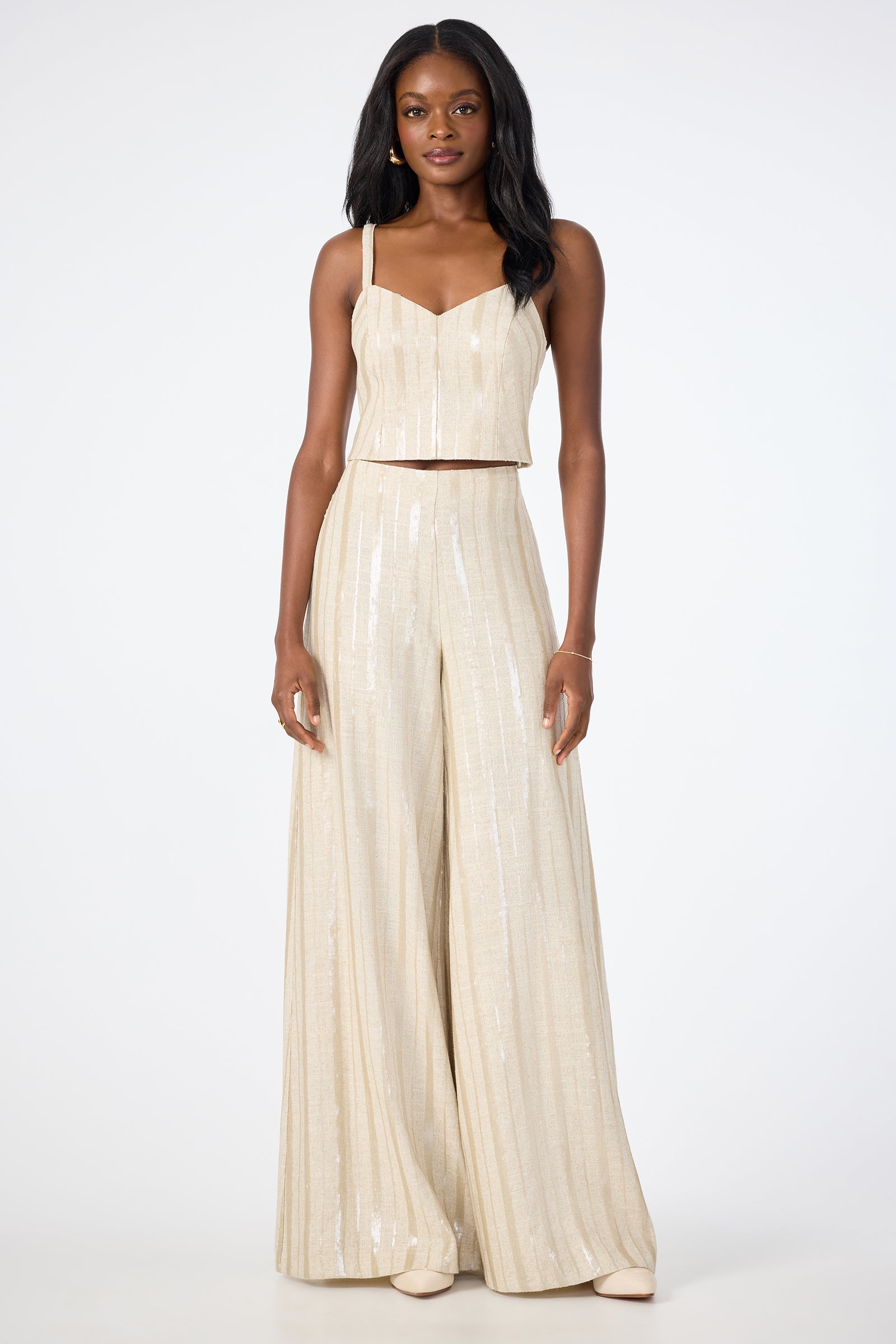 A woman faces forward wearing Nice Wide-Leg Pants—a cream, sleeveless two-piece with subtle vertical stripes and a shimmery finish. The top is cropped, and the wide-leg pants are floor-length. She completes the look with nude shoes.