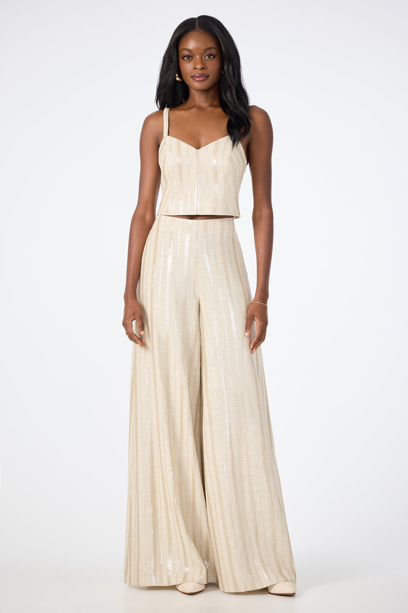 A woman faces forward in the Millau Sleeveless Top, styled with a light gold, sequin wide-leg jumpsuit featuring thin straps and paired with nude shoes, set against a plain white background.