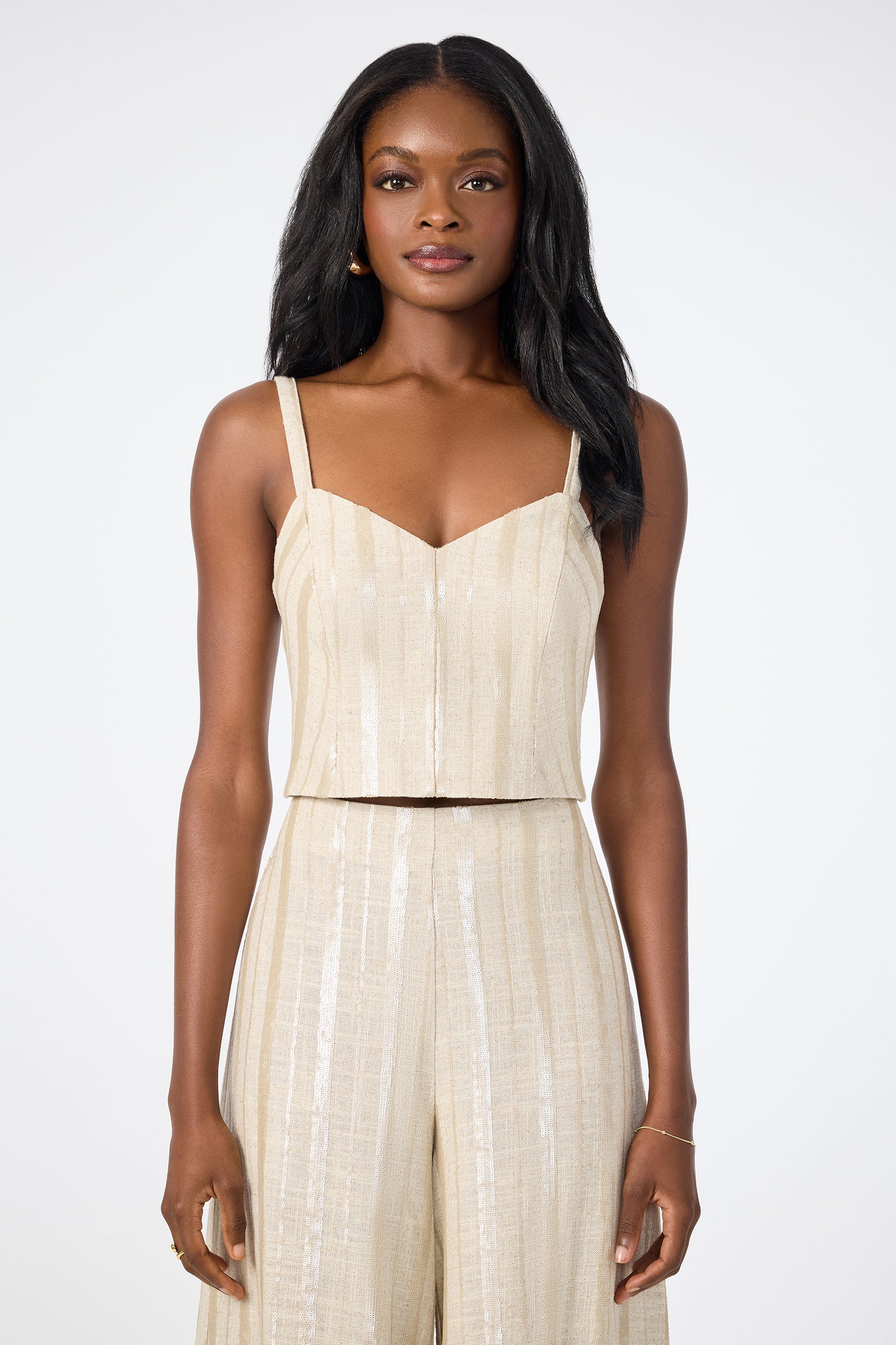 A woman with long, dark hair wears the Millau Sleeveless Top—beige, textured, and cropped—paired with matching high-waisted pants while standing against a plain white background.