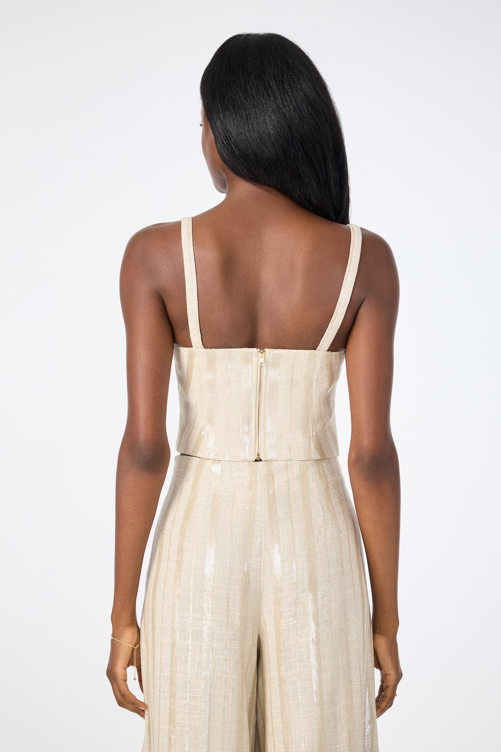 A woman with long dark hair is shown from behind, wearing the Millau Sleeveless Top in light gold. The shimmery top features a back zipper and is styled against a plain white background.