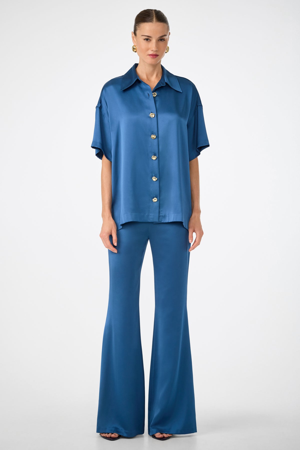 A person faces forward in a loose, blue satin Manju Button-Front Top with matching flared pants. The outfit is short-sleeved, their hair is pulled back, and the background is plain white.