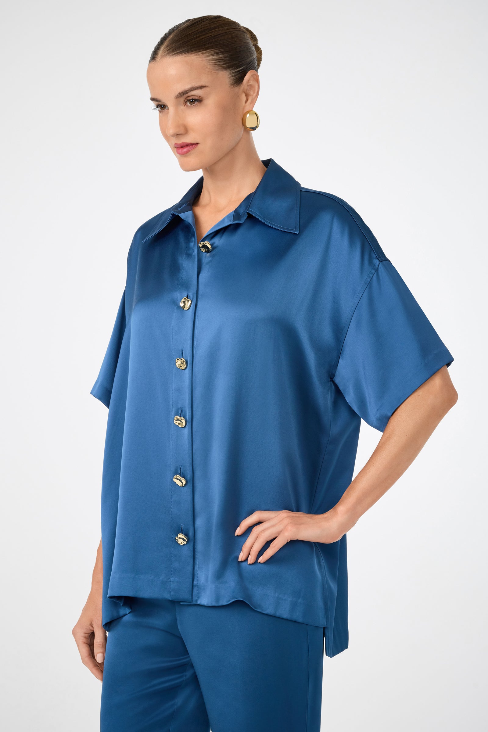 A woman wears the Manju Button-Front Top, a shiny, loose-fitting blue satin short-sleeve shirt, paired with matching blue pants, standing with one hand on her hip against a plain light background.
