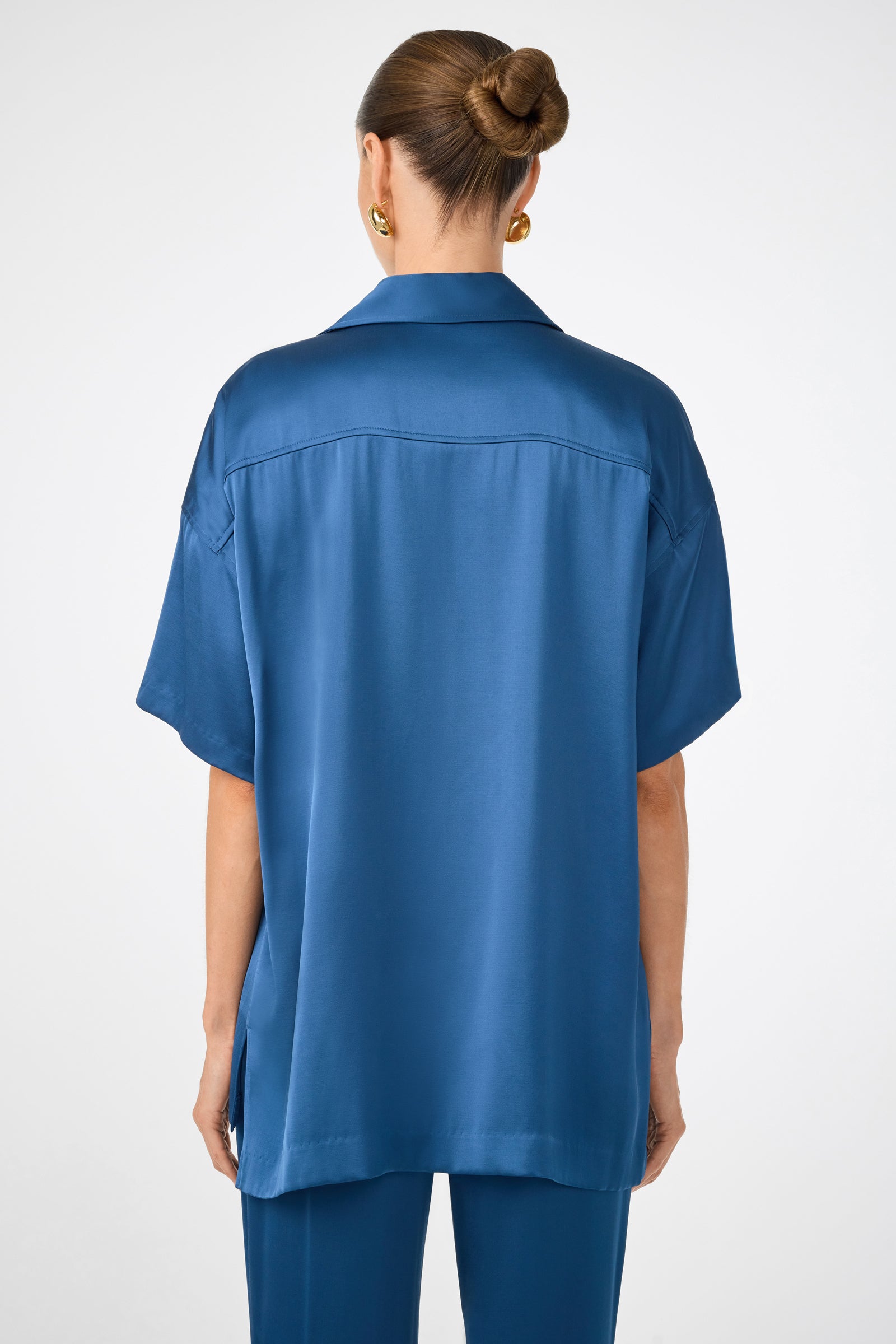 A woman with hair in a bun and gold earrings is seen from the back, modeling the Manju Button-Front Top—a loose-fitting, short-sleeved blue satin shirt with matching pants—against a plain light background.