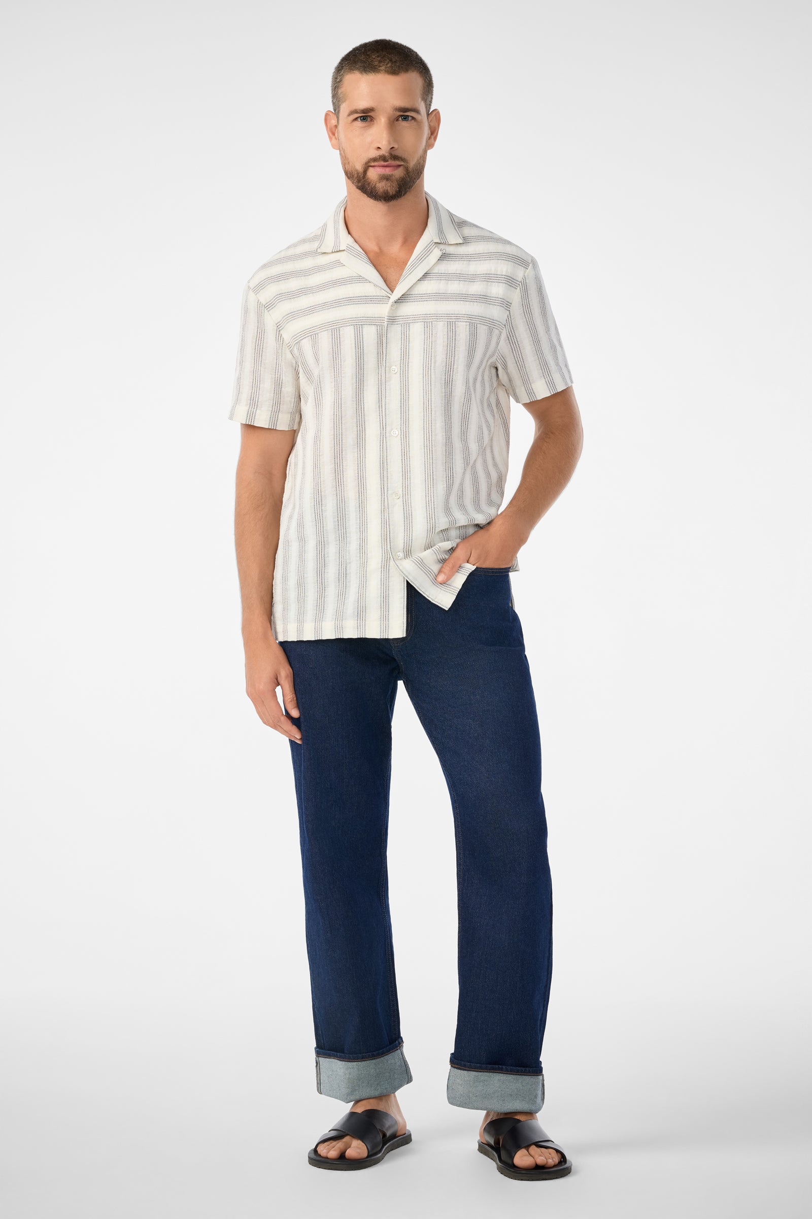 A man with short hair and a beard wears the Angelo Short-Sleeve Shirt - Stripe Gauze, paired with dark cuffed jeans and black sandals, standing with one hand in his pocket against a plain white background.