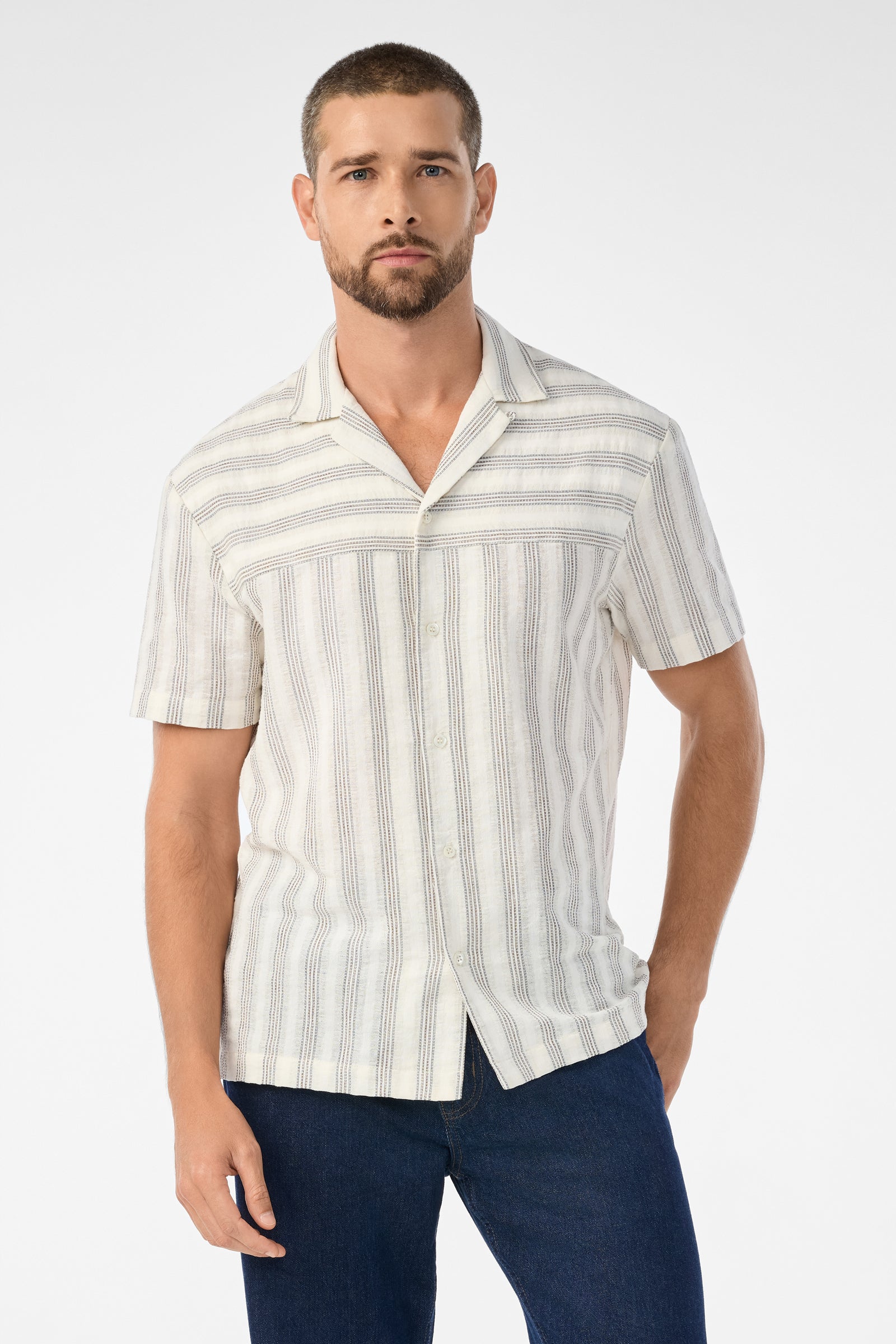 A man with short hair and a trimmed beard wears the Angelo Short-Sleeve Shirt - Stripe Gauze in Ecru, paired with dark blue jeans. He stands against a plain white background.