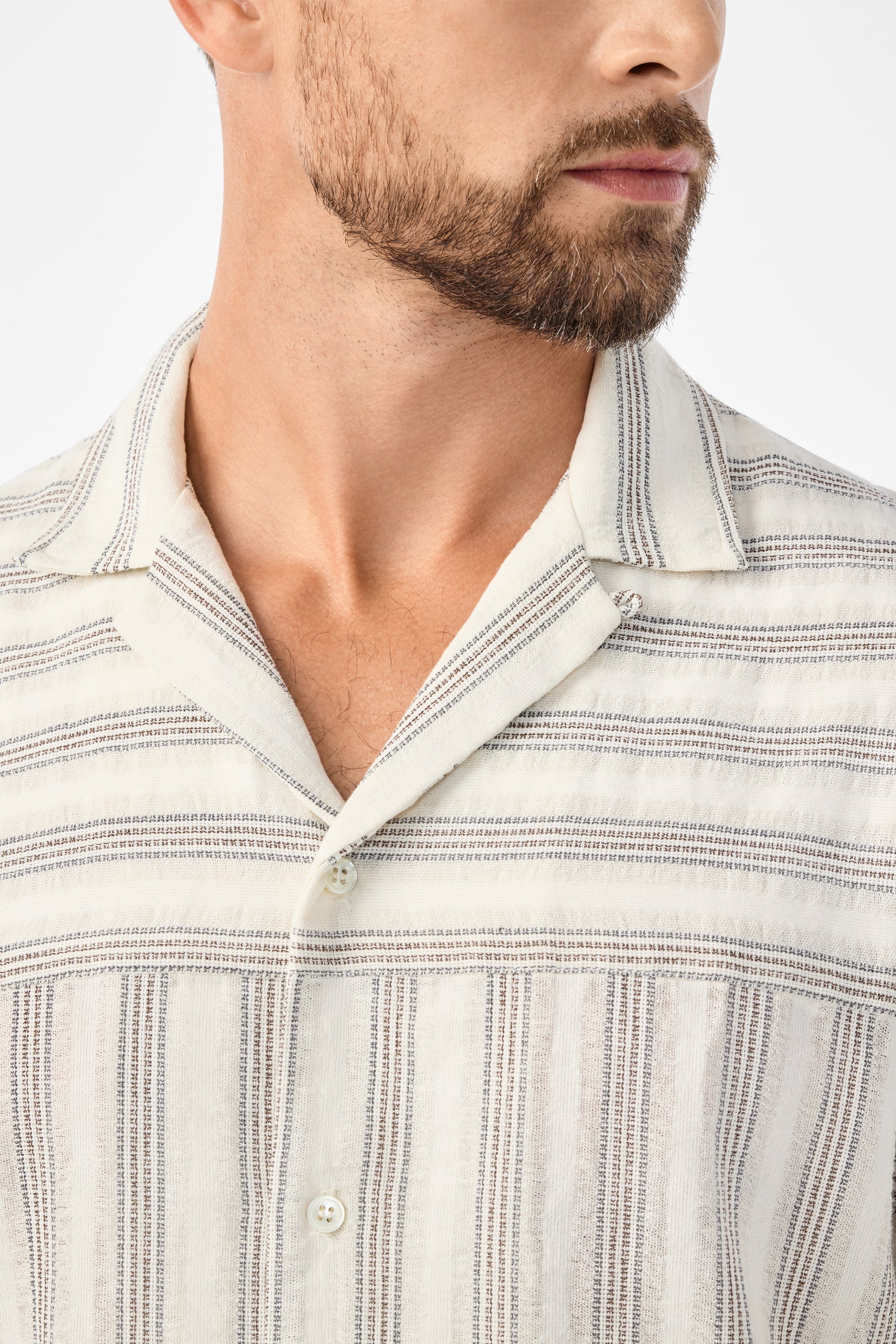 Close-up of a man in the Angelo Short-Sleeve Shirt - Stripe Gauze: a cream-colored, short-sleeve shirt with thin grey stripes and a camp collar. His trimmed beard is visible against a plain white background.