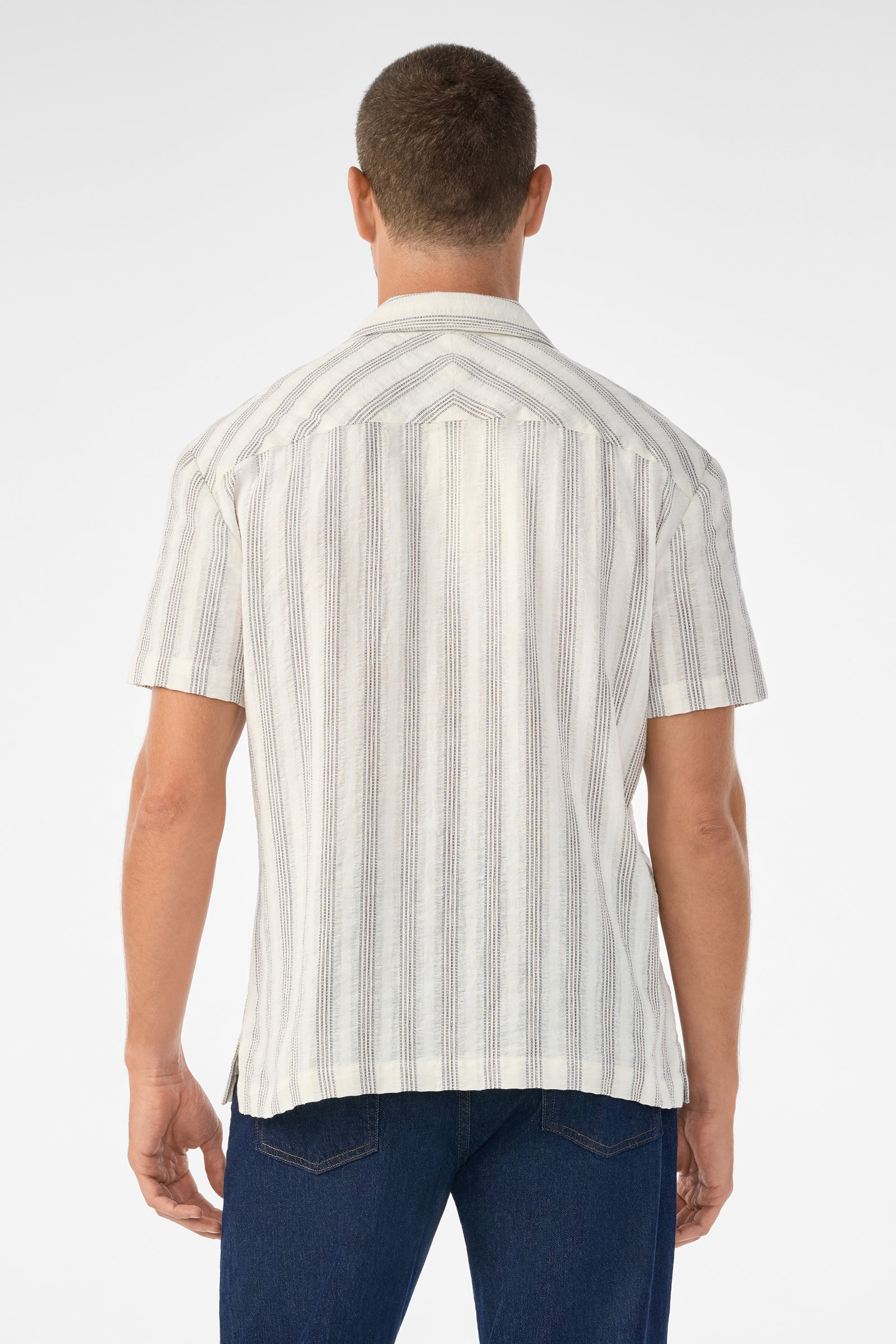 A man is seen from behind wearing the Angelo Short-Sleeve Shirt - Stripe Gauze, a white button-up with thin vertical stripes, paired with blue jeans against a plain white background.