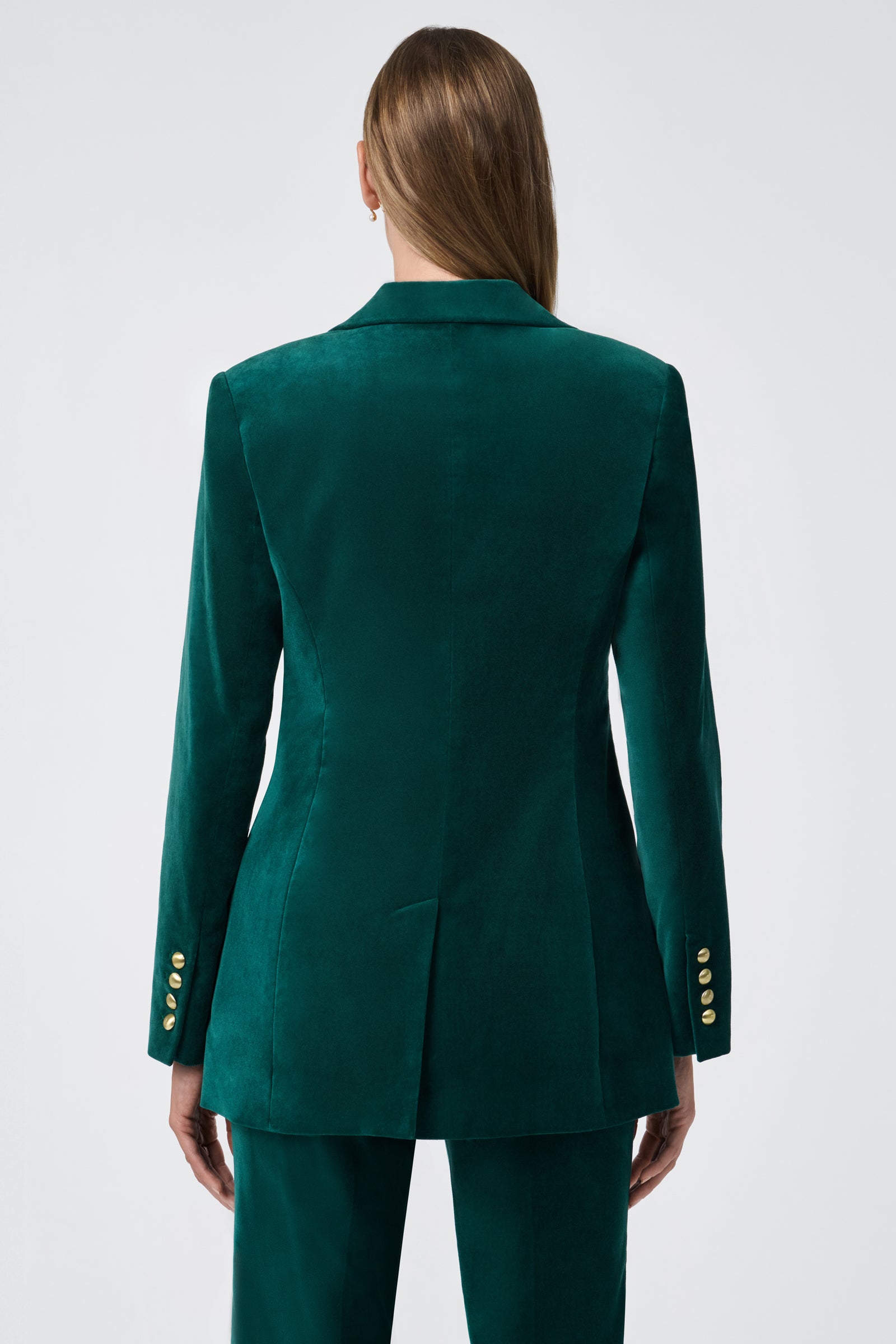 A person with straight, long light brown hair wears the Broadway Jacket—a teal velvet blazer with gold sleeve buttons and a center seam—paired with matching trousers. Shown from the back against a plain, light background.