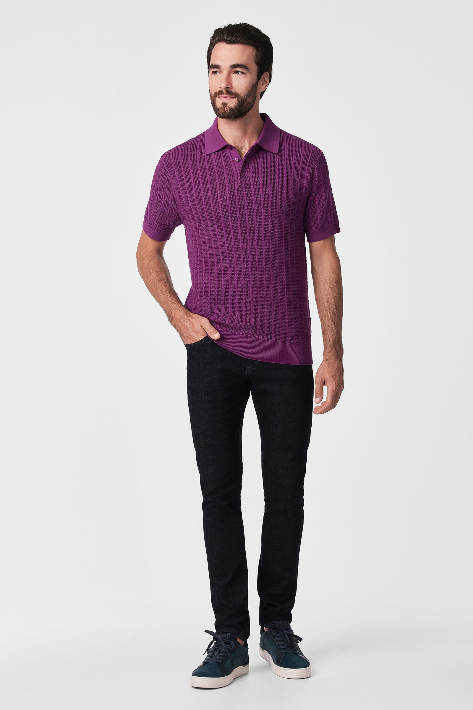 A man with short dark hair and a beard wears the Asher Ss Polo, a purple short-sleeve knit shirt, paired with black jeans and dark sneakers. He stands against a plain white background, looking slightly right with one hand in his pocket.