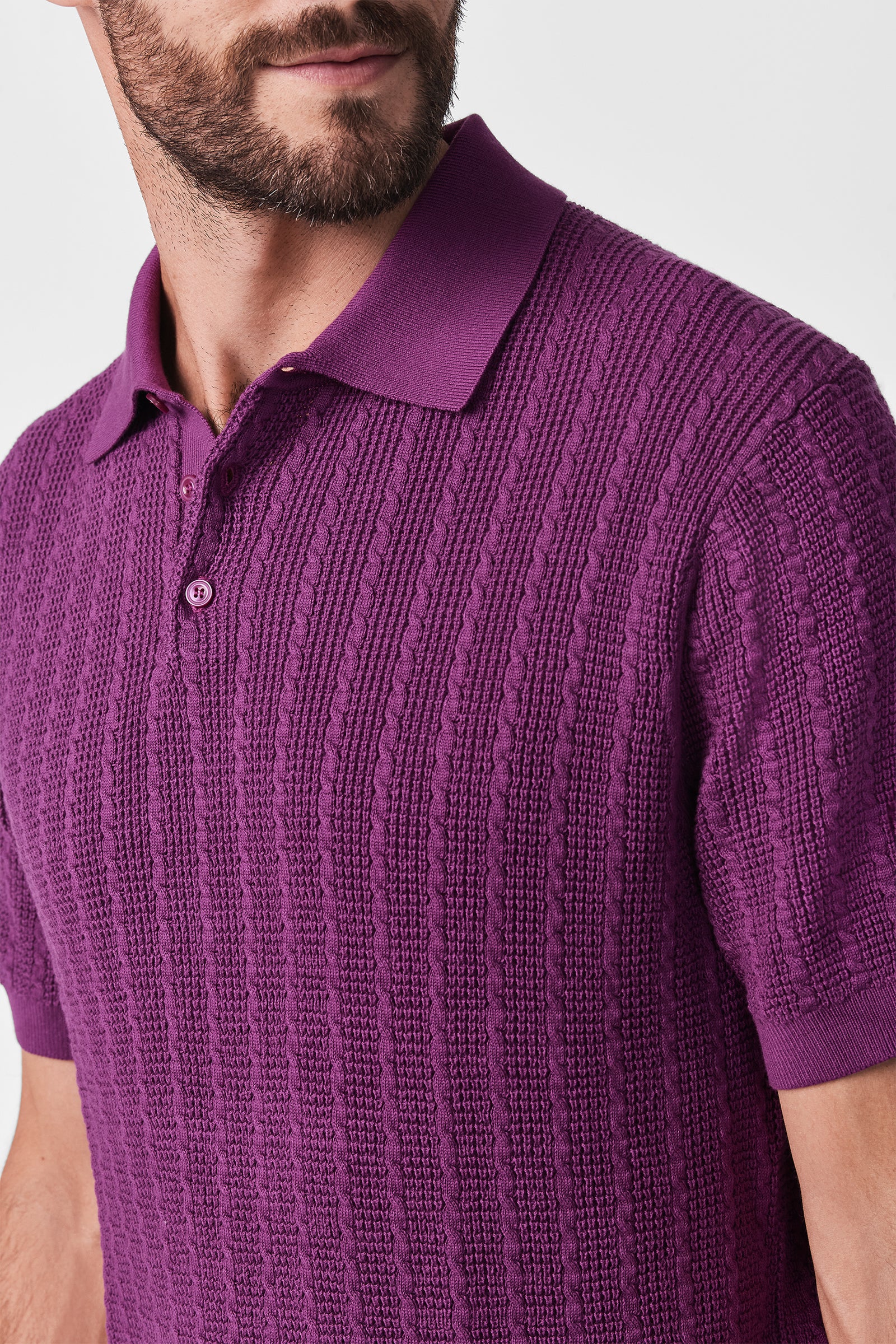 A man with a beard wears the Asher Ss Polo, a textured, short-sleeve purple shirt with a collar and buttons. The cropped image shows his upper torso and part of his face against a plain white background.