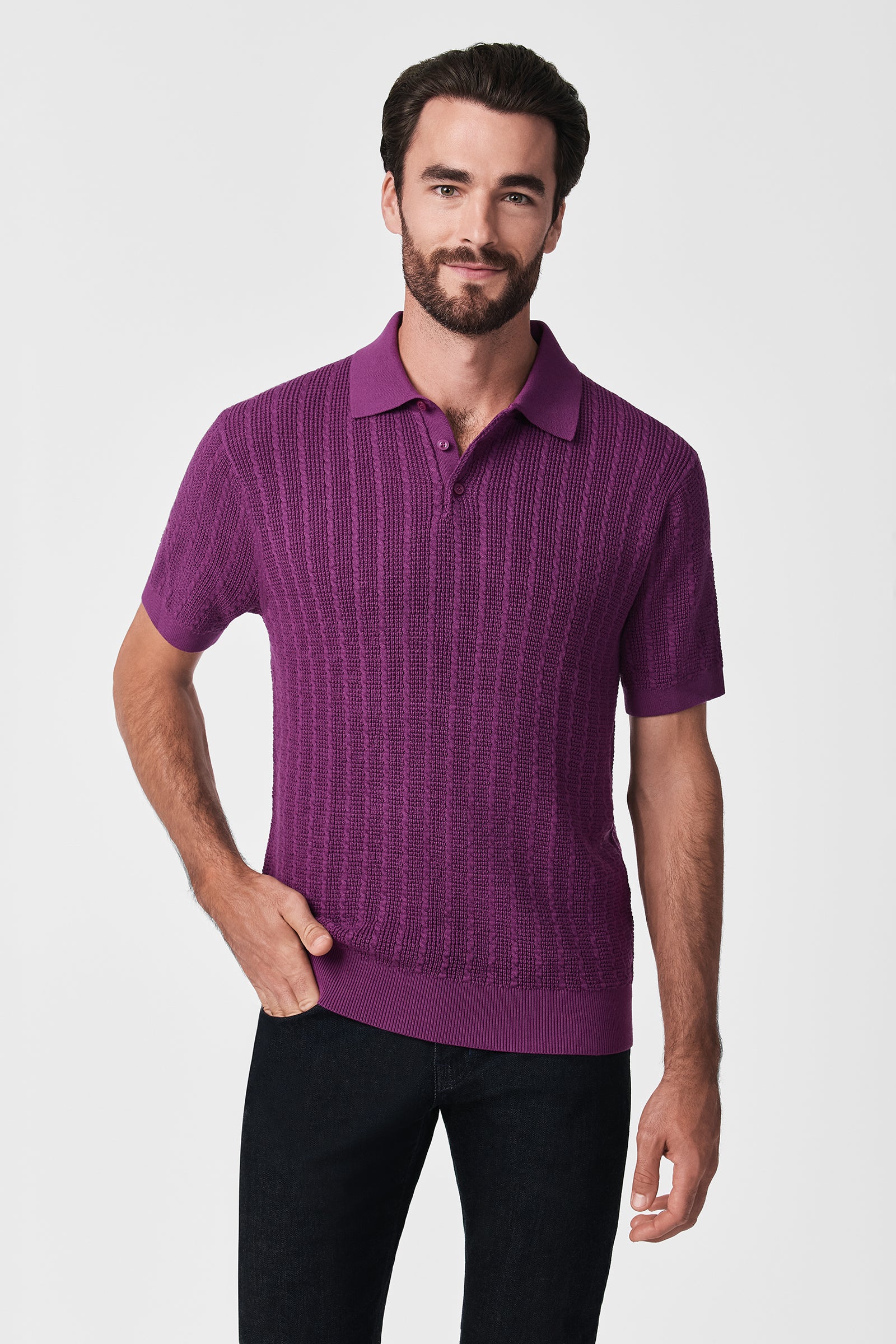 A man with short dark hair and a beard wears the Asher Ss Polo, a textured short-sleeve purple shirt, paired with black pants and stands with one hand in his pocket against a plain white background.