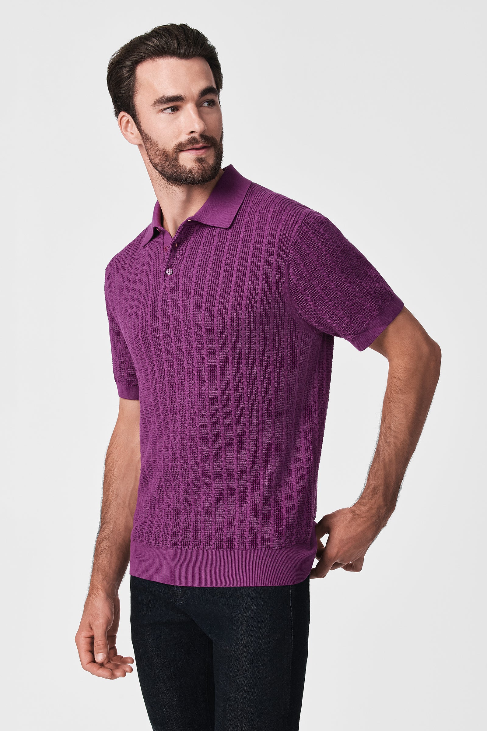 A man with dark hair and a beard wears the Asher Ss Polo—a textured, short-sleeve purple shirt—and dark pants, standing against a plain light background while looking to his left.