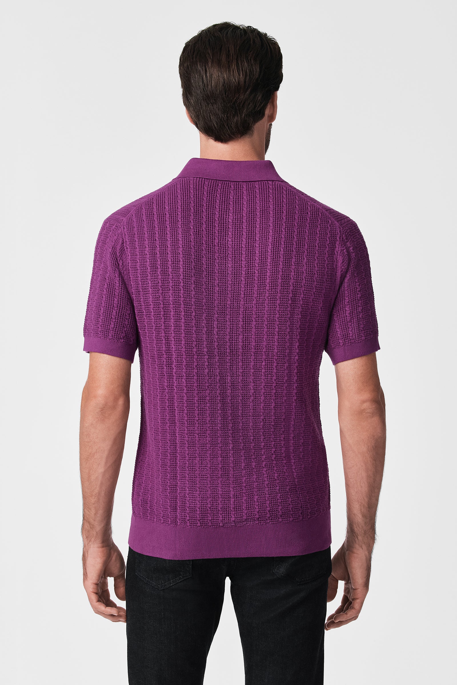 A man faces away, wearing the Asher Ss Polo—a textured, short-sleeved purple knit shirt—with black pants, set against a plain white background.