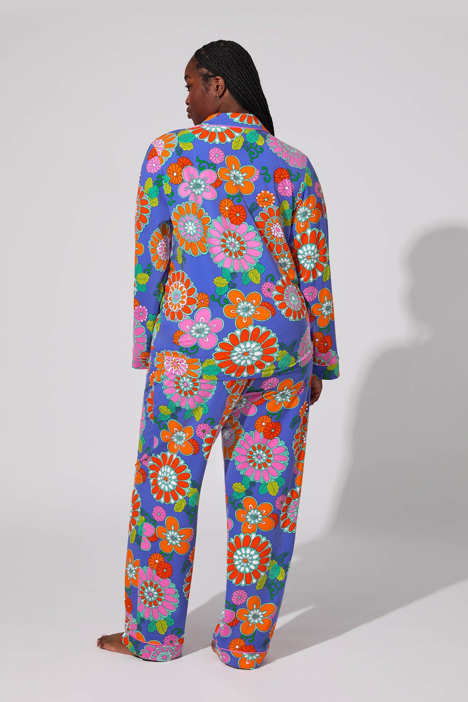 A person stands barefoot facing away, wearing the Classic PJ Set - Tokyo Floral, featuring bright oversized orange, pink, green, and white floral patterns on a purple background against a plain light backdrop.