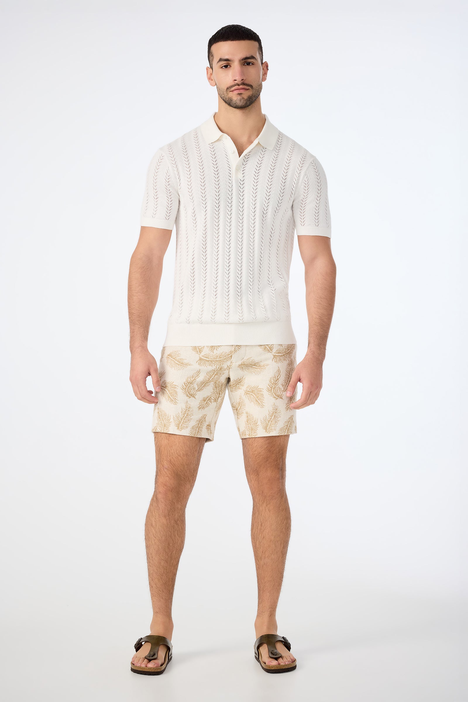 A man stands against a plain white background wearing the Cole Short-Sleeve Polo in white, beige leaf-patterned shorts, and brown sandals. He faces forward with a neutral expression and relaxed posture.