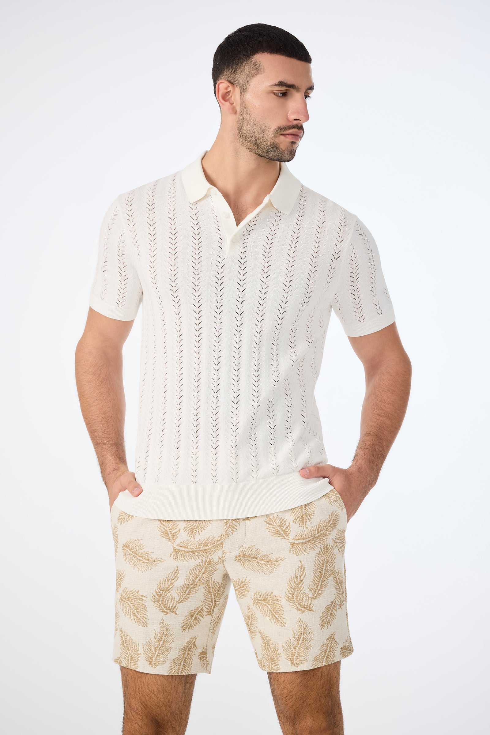 A man wears the Cole Short-Sleeve Polo in Whitewash with a delicate leaf pattern and matching beige shorts, standing with his hands in his pockets against a plain white background.