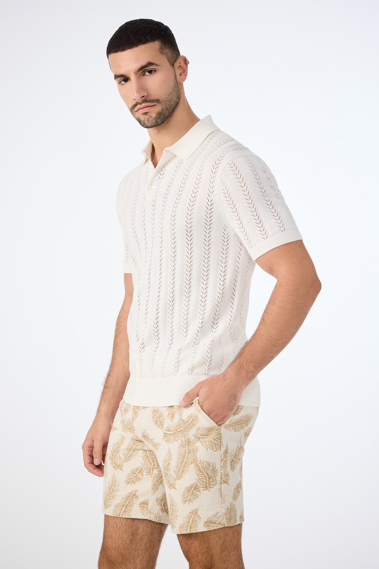 A man with short dark hair and a trimmed beard models the Cole Short-Sleeve Polo in white with a subtle pattern, paired with beige leaf-print shorts, posing against a plain light background with one hand in his pocket.