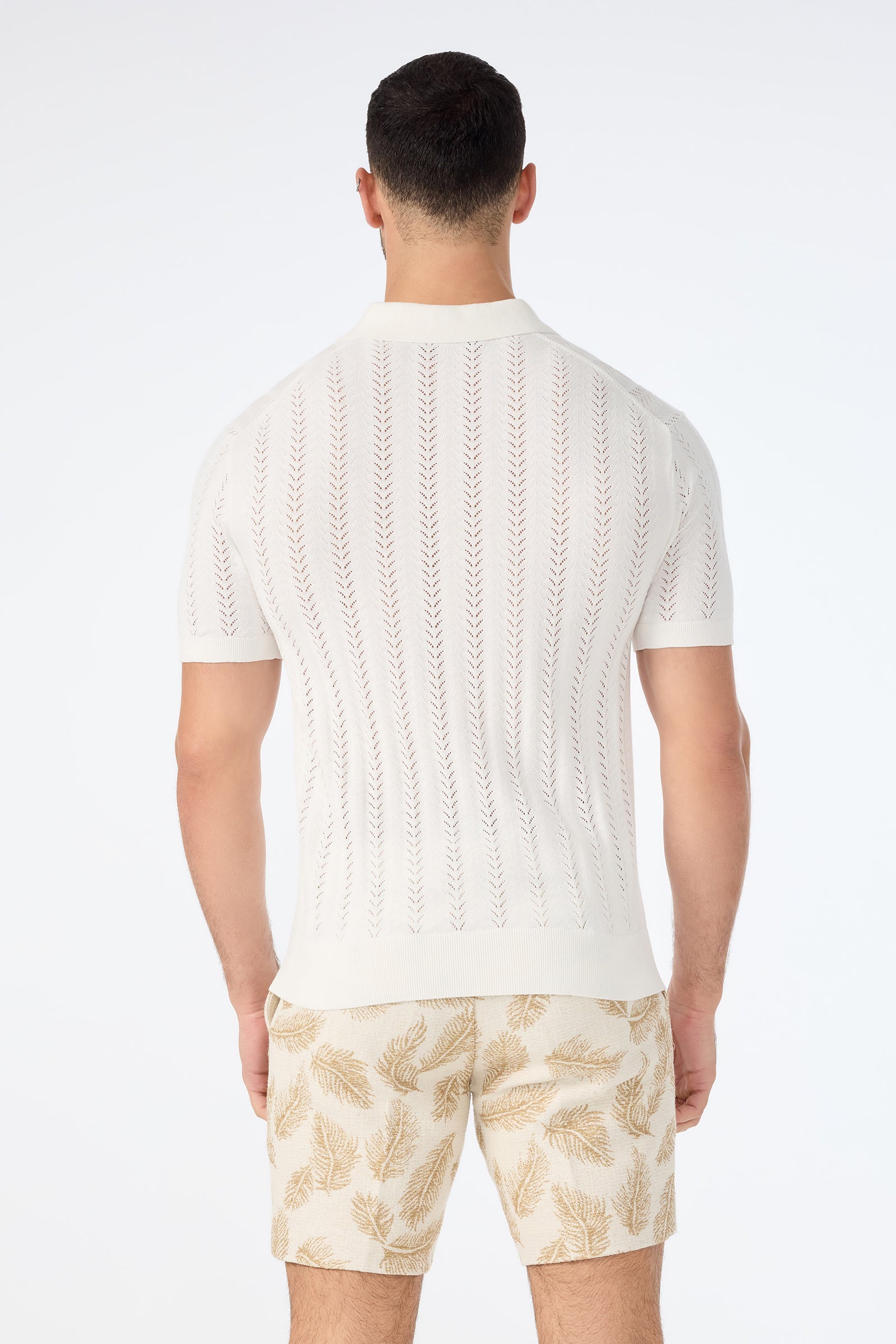 A man faces away, wearing the Cole Short-Sleeve Polo in white with a vertical pattern and beige leafy-print shorts. The background is plain white.