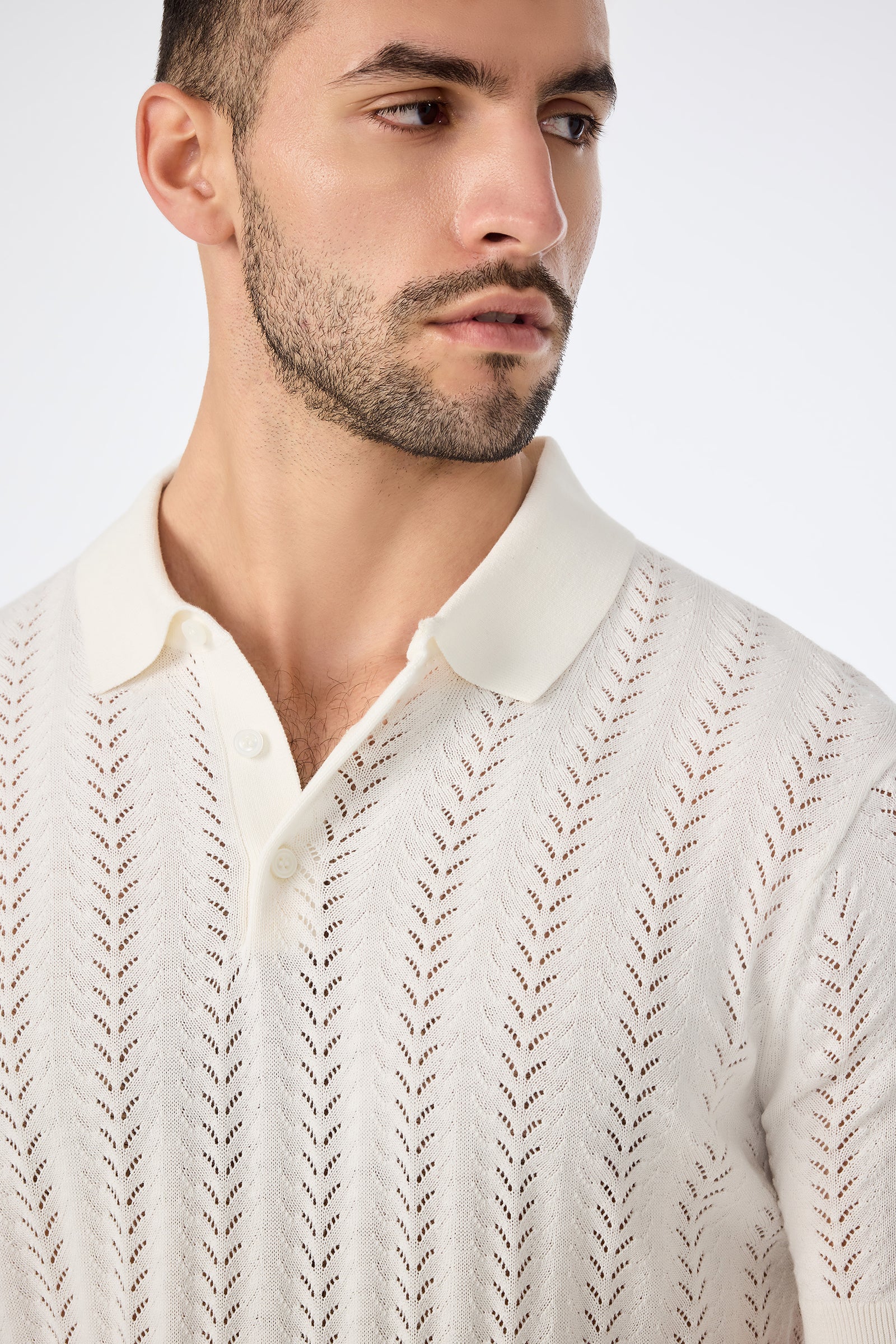 A man with short dark hair and a trimmed beard models the Cole Short-Sleeve Polo, a cream-colored collared knit shirt with a chevron lace pattern. He looks to the side against a plain light background.