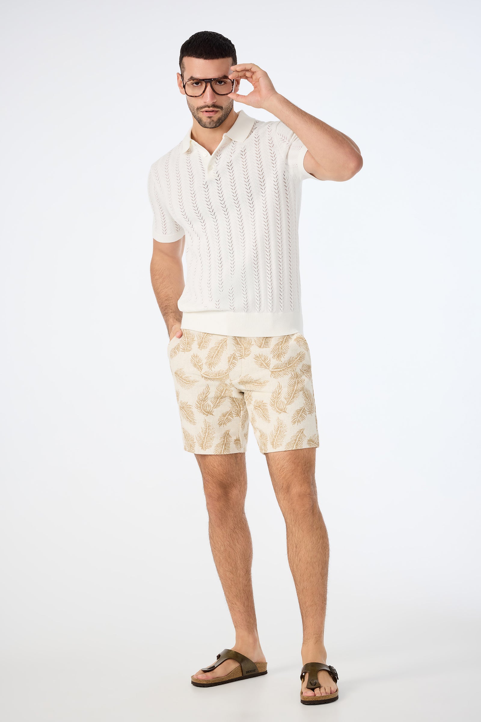 A man wearing glasses, a white Cole Short-Sleeve Polo, beige leaf-patterned shorts, and brown sandals stands against a plain white background, looking at the camera with one hand touching his glasses.