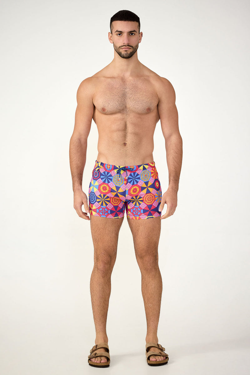 A man stands facing forward against a plain background, wearing the Palm Desert Swim Trunk - Parasol and brown sandals. He is shirtless with a neutral expression.