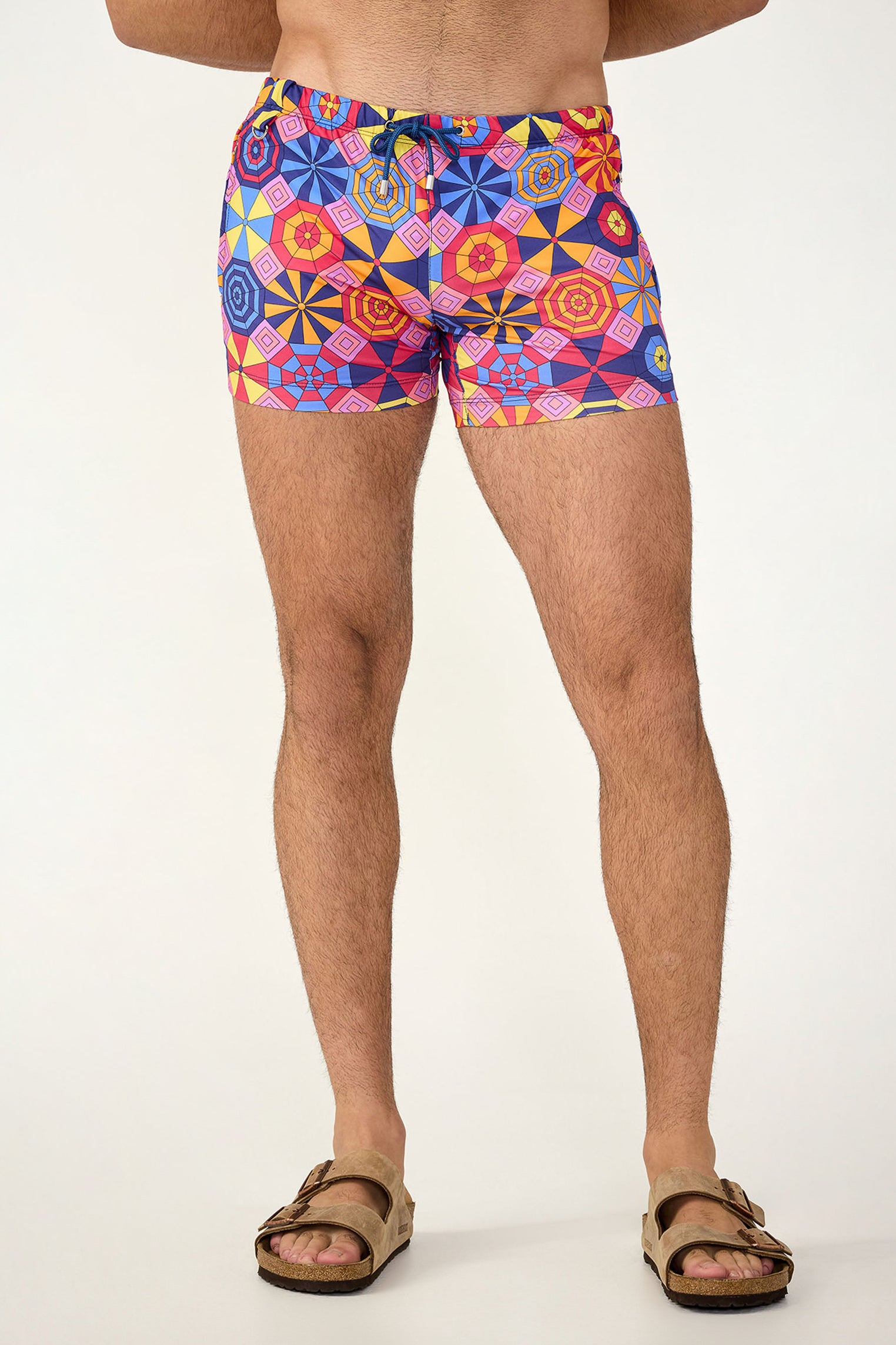 A person, shown from the waist down against a plain light background, wears the Palm Desert Swim Trunk - Parasol in Multi along with brown sandals.