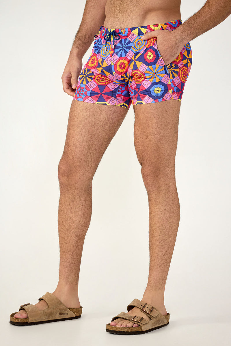 A person stands sideways against a plain background, wearing the Palm Desert Swim Trunk - Parasol featuring colorful geometric patterns in blue, pink, orange, and yellow, paired with beige slide sandals.