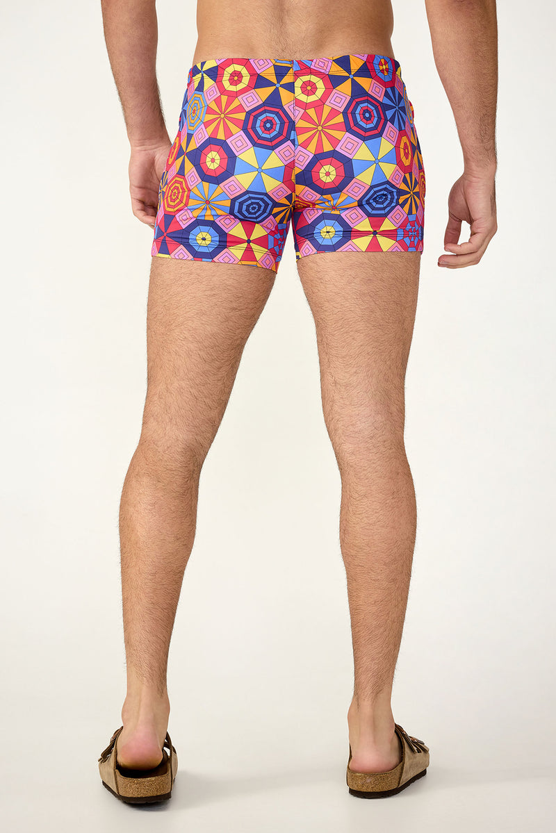 A man stands facing away, wearing the Palm Desert Swim Trunk - Parasol with colorful geometric patterns and tan sandals. The image highlights his legs and back against a plain, light background.