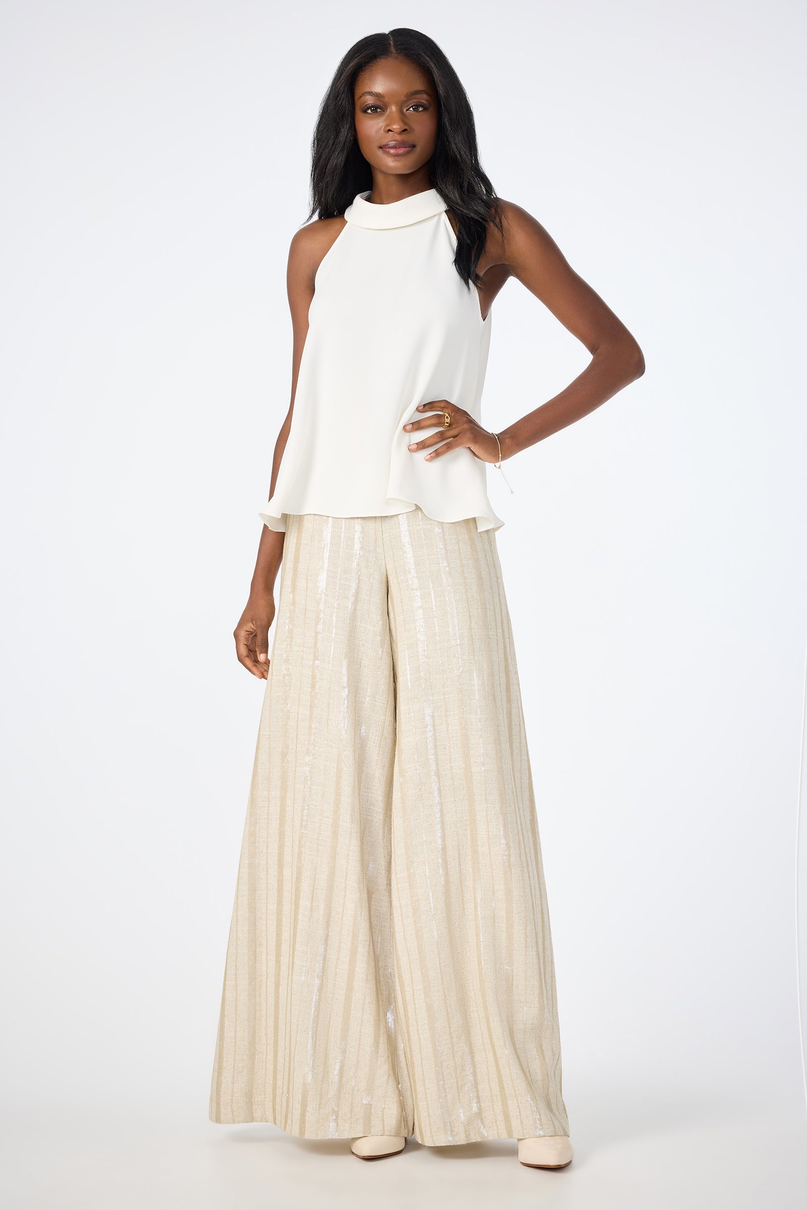 A woman stands confidently in the Eze Halter Top, a white sleeveless piece, paired with high-waisted wide-leg beige pants featuring a subtle shimmer. She has long dark hair and light shoes, posed against a plain white background.