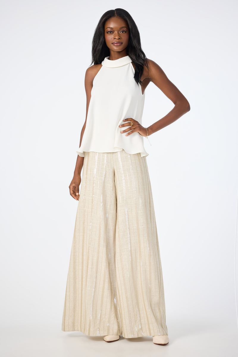 A woman stands confidently in the Eze Halter Top, a white sleeveless piece, paired with high-waisted wide-leg beige pants featuring a subtle shimmer. She has long dark hair and light shoes, posed against a plain white background.