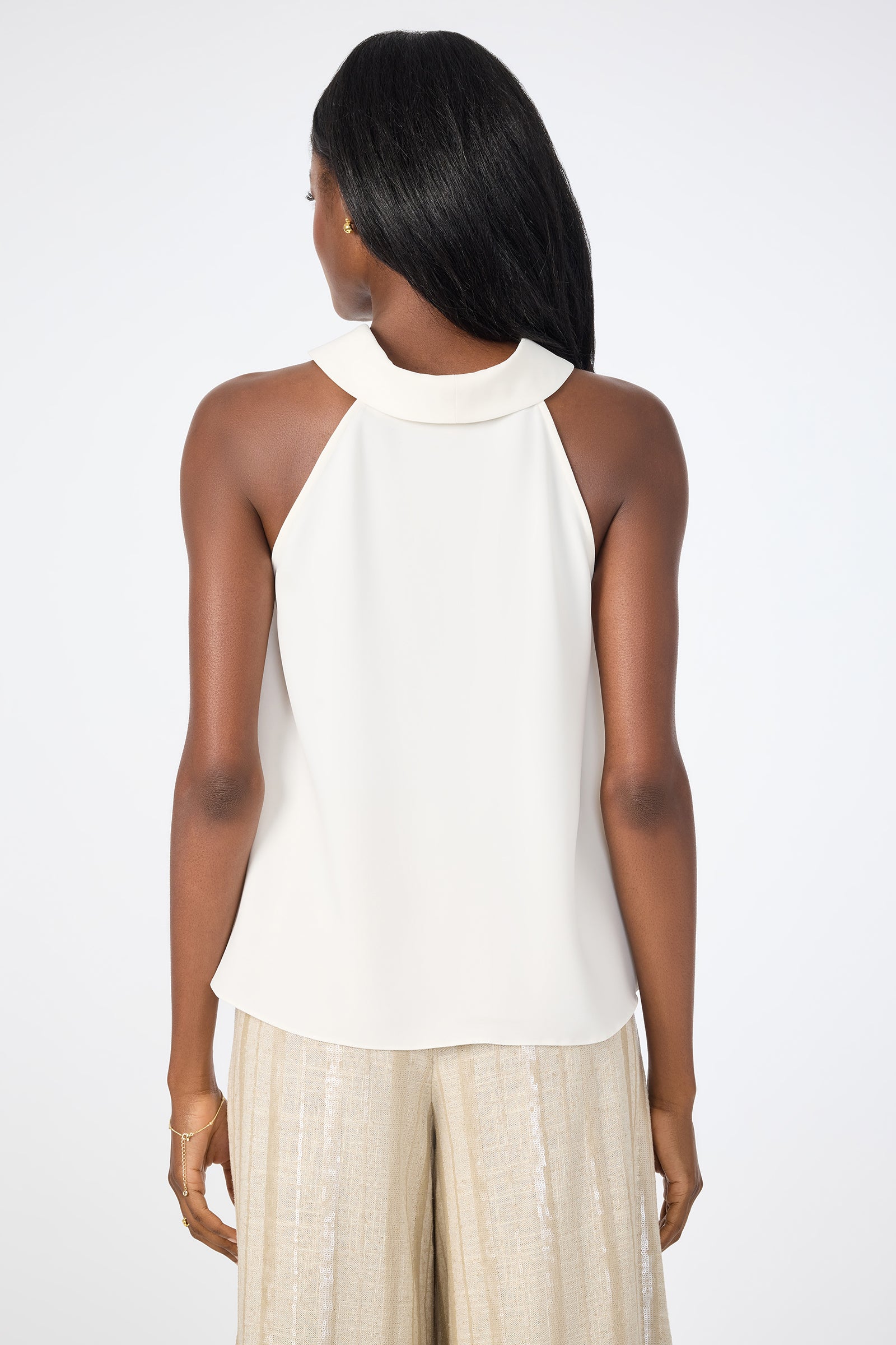 A woman with long, straight black hair and minimal jewelry stands with her back to the camera, wearing the sleeveless white Eze Halter Top and light beige pleated pants against a plain white background.
