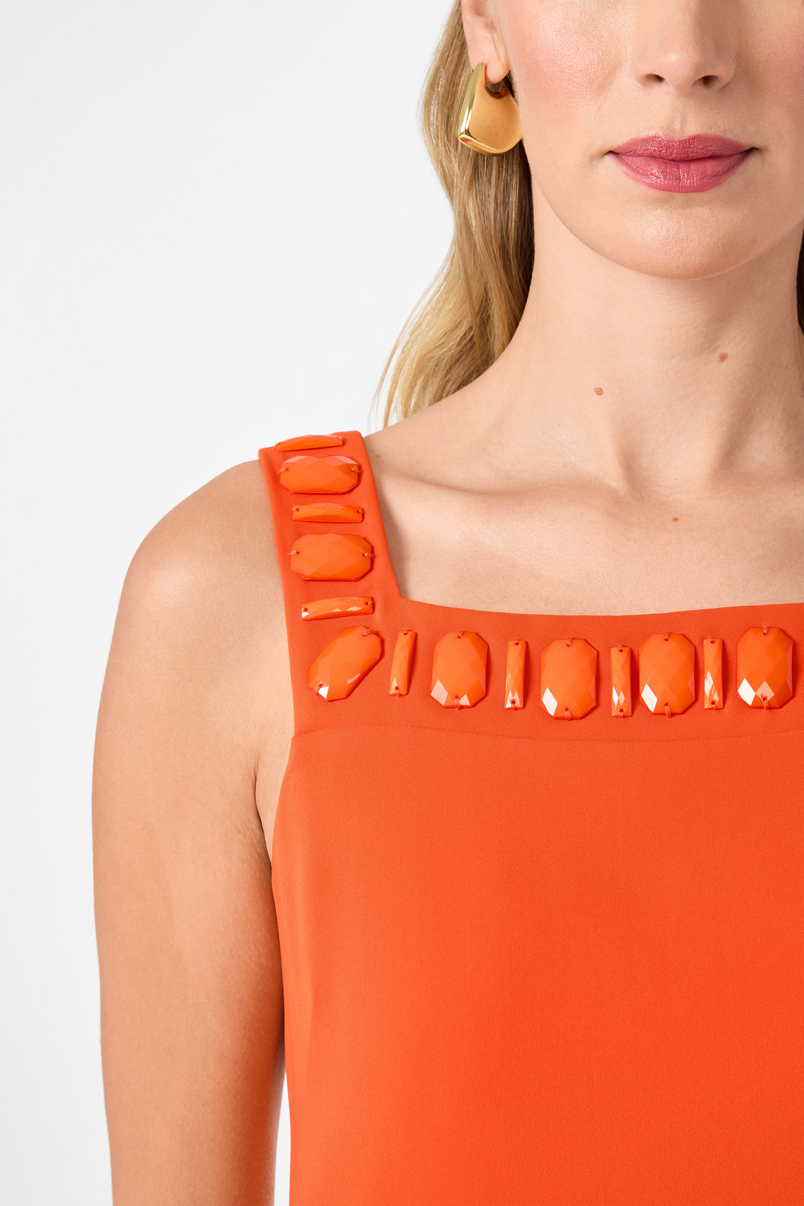A woman wears the Yokuna Jewel Neck Mini Dress, a bright orange sleeveless style with large, shiny embellishments at the neckline. She has wavy blonde hair, gold hoop earrings, and pink lipstick against a plain white background.