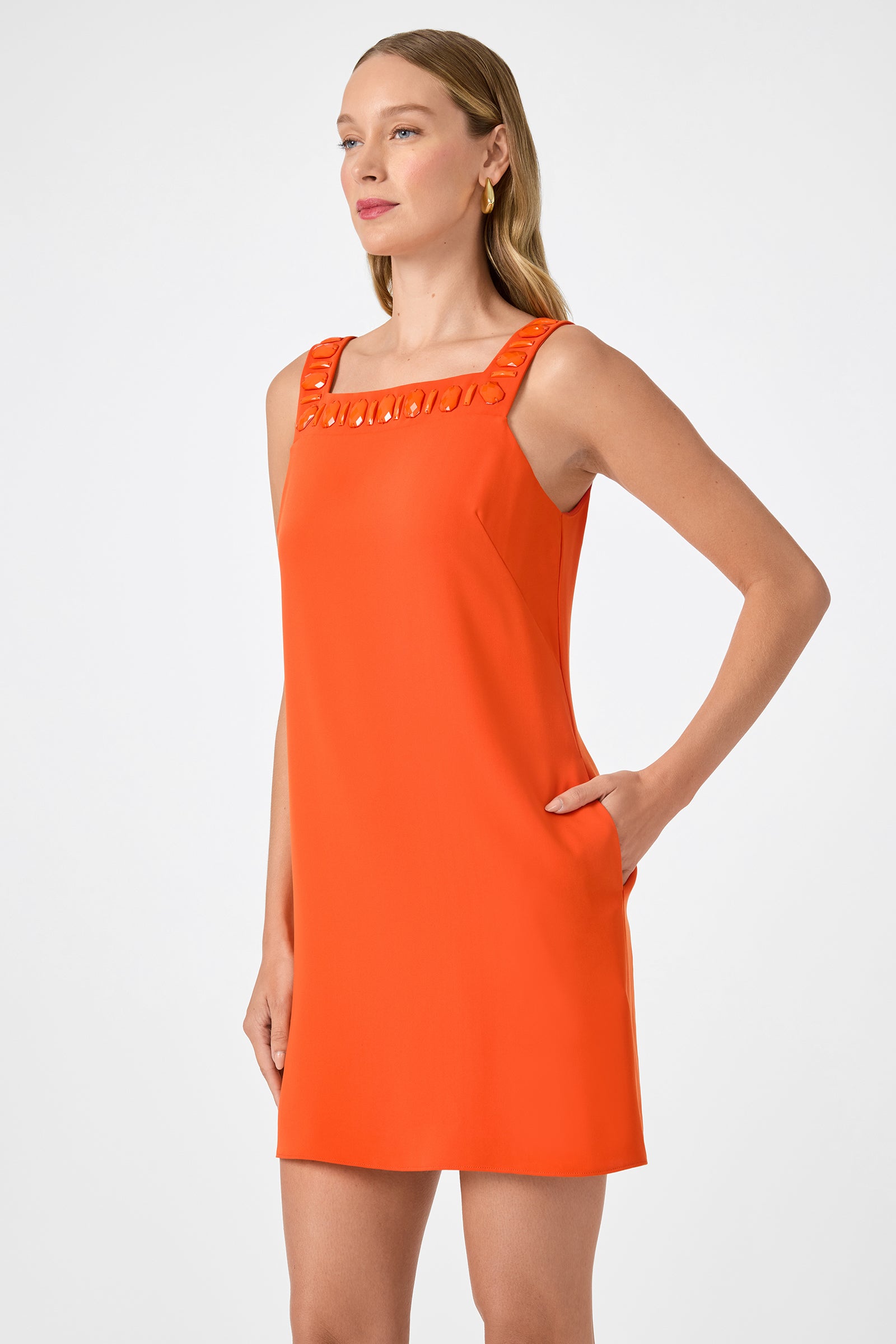 A woman stands confidently with one hand in her pocket, wearing the Yokuna Jewel Neck Mini Dress—a sleeveless, bright orange shift featuring a square neckline and decorative accents along the neckline.