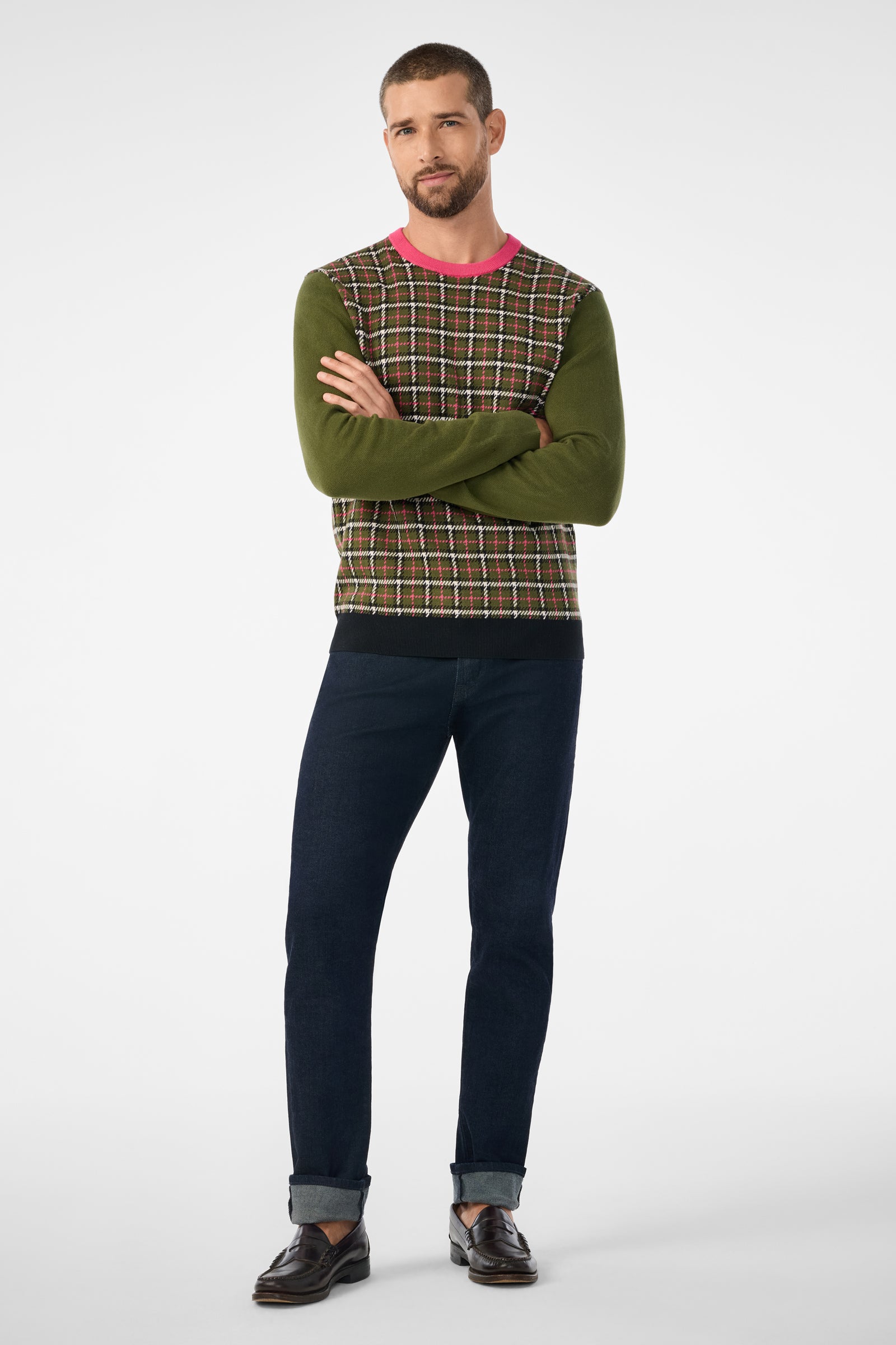 A man stands with arms crossed, wearing the Buchanan Long-Sleeve Crewneck sweater in green and brown plaid with a pink collar, dark blue cuffed jeans, and brown loafers against a plain light background.
