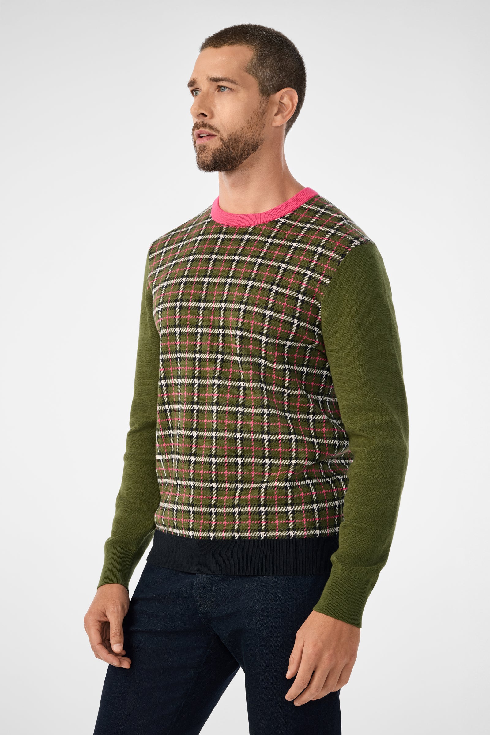 A man with short hair and a beard wears the Buchanan Long-Sleeve Crewneck, featuring a plaid pattern with green sleeves and a bright pink collar, paired with dark pants against a plain light background.