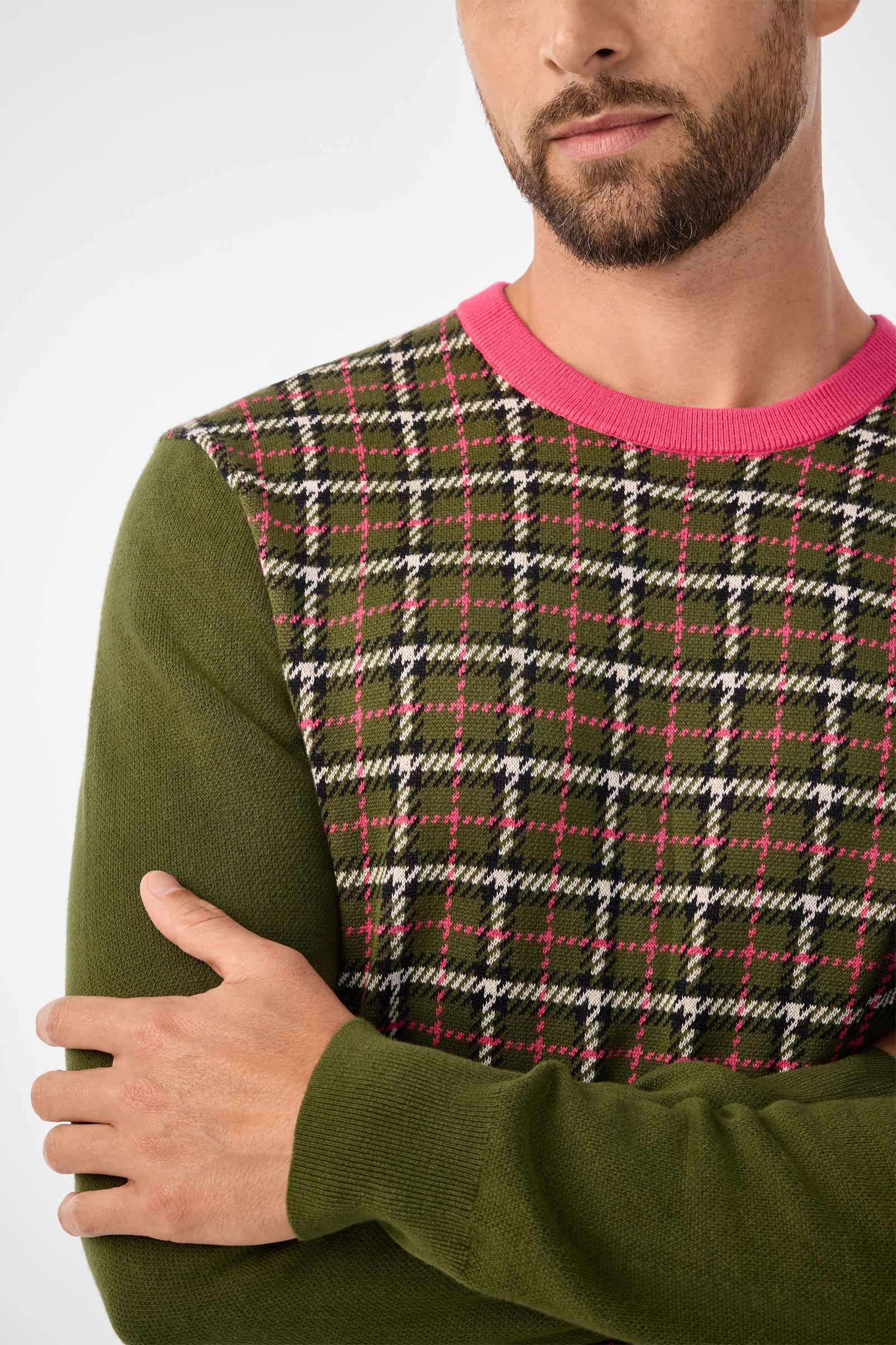 A man with a short beard crosses his arms, wearing the Buchanan Long-Sleeve Crewneck—a green sweater with a green, pink, black, and white plaid front panel and a pink collar. The photo is cropped at the nose and above the waist.