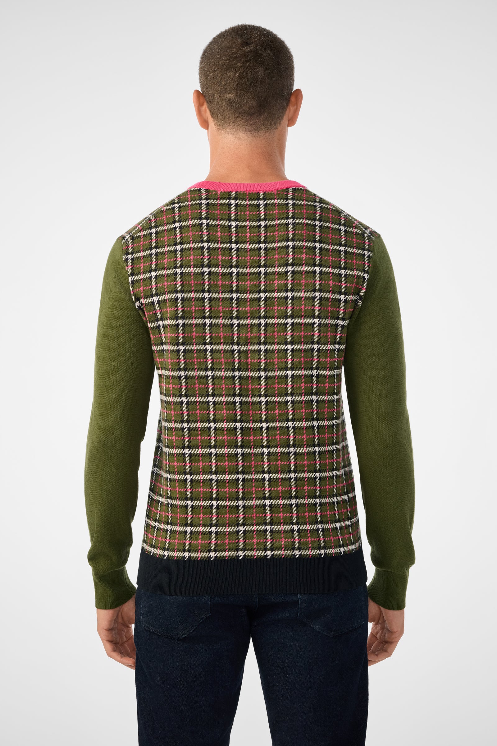 A person with short hair is seen from behind, wearing the Buchanan Long-Sleeve Crewneck with green sleeves, a pink collar, dark cuffs, and a plaid-patterned back in brown, pink, white, and black. They are also wearing dark pants.