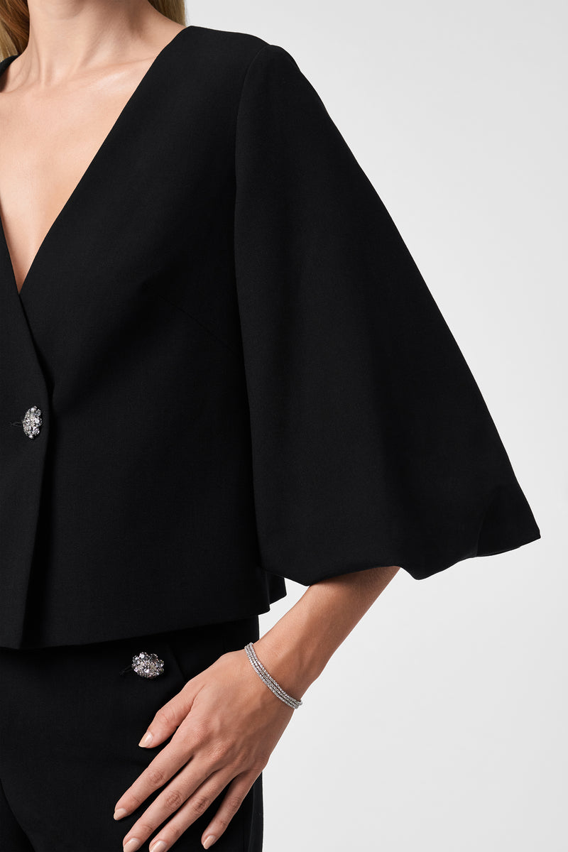 A woman wears the Basal Cropped Jacket, a black piece with wide, flowing sleeves and jeweled buttons, paired with a thin silver bracelet. The image is cropped from shoulders to hips against a plain background.