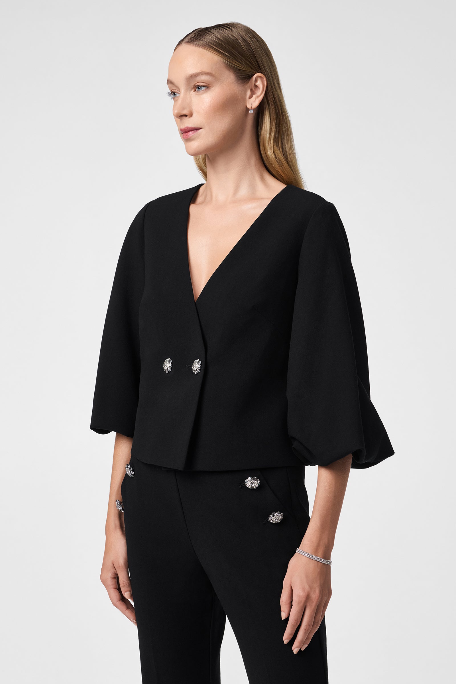 A woman with long, blonde hair wears the Basal Cropped Jacket—a black V-neck top with wide 3/4 sleeves and jeweled buttons—paired with matching black pants featuring jeweled buttons, against a plain light background.
