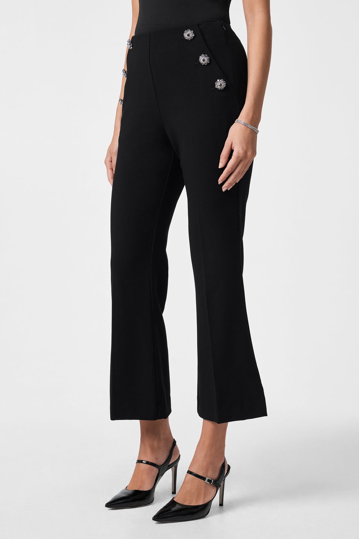 A woman stands against a white background wearing the Luhrmann Pant—black, high-waisted, slightly flared with three decorative waist buttons—paired with a black sleeveless top, black heels, and silver bracelets.