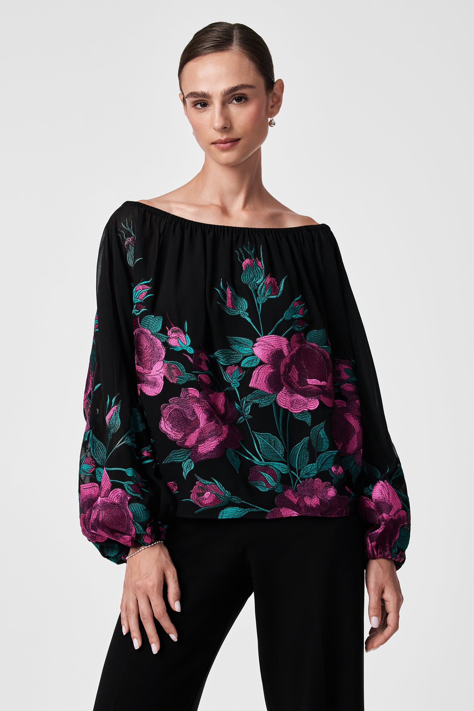 A woman stands against a plain light background wearing the Soho 2 Top—an off-the-shoulder black blouse with voluminous sleeves and a vibrant pink and green floral print—paired with black pants.