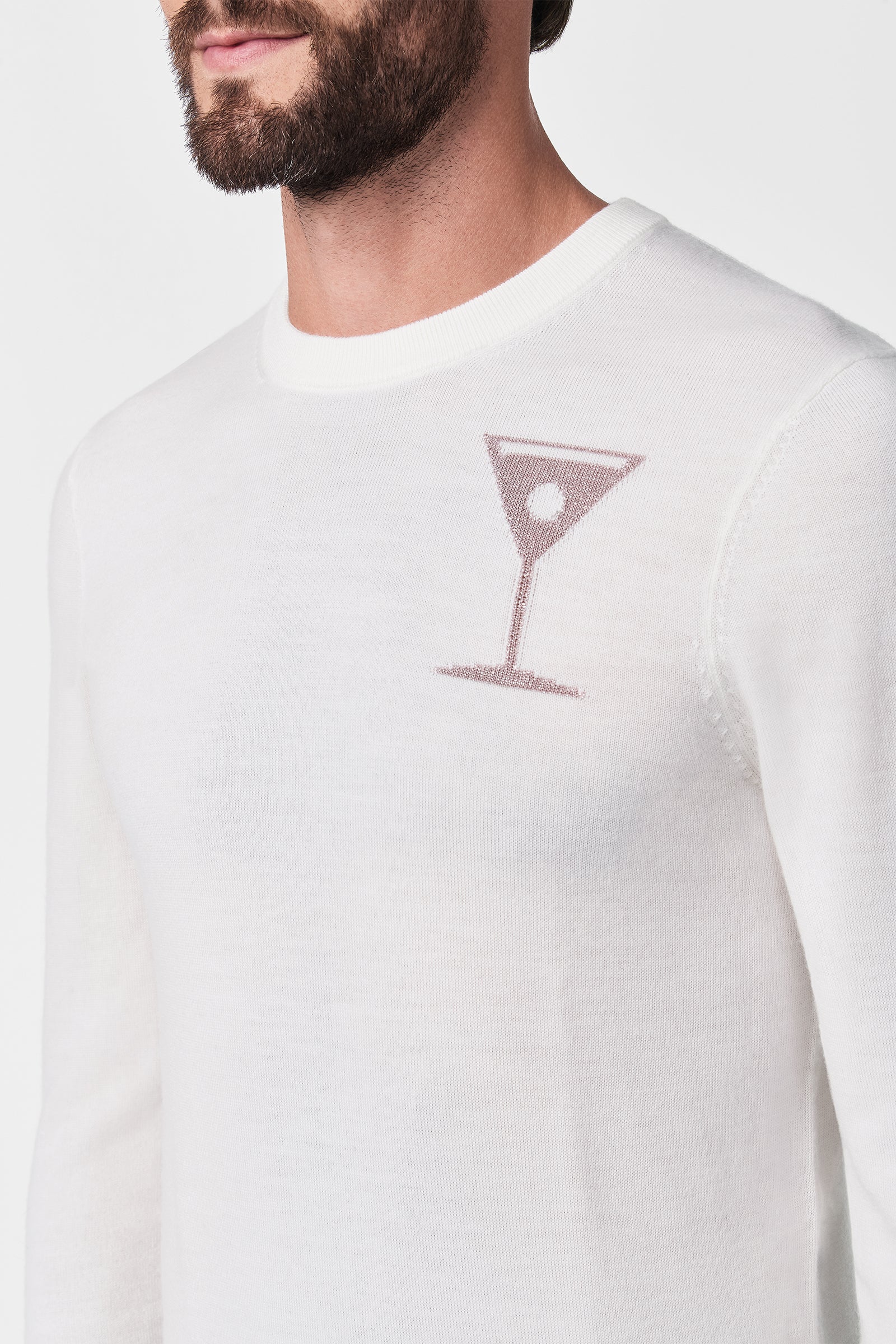 A man wearing the Martini Crewneck, a white sweater featuring a small, minimalist martini glass outlined in light brown with an olive on the chest. The background is plain white.