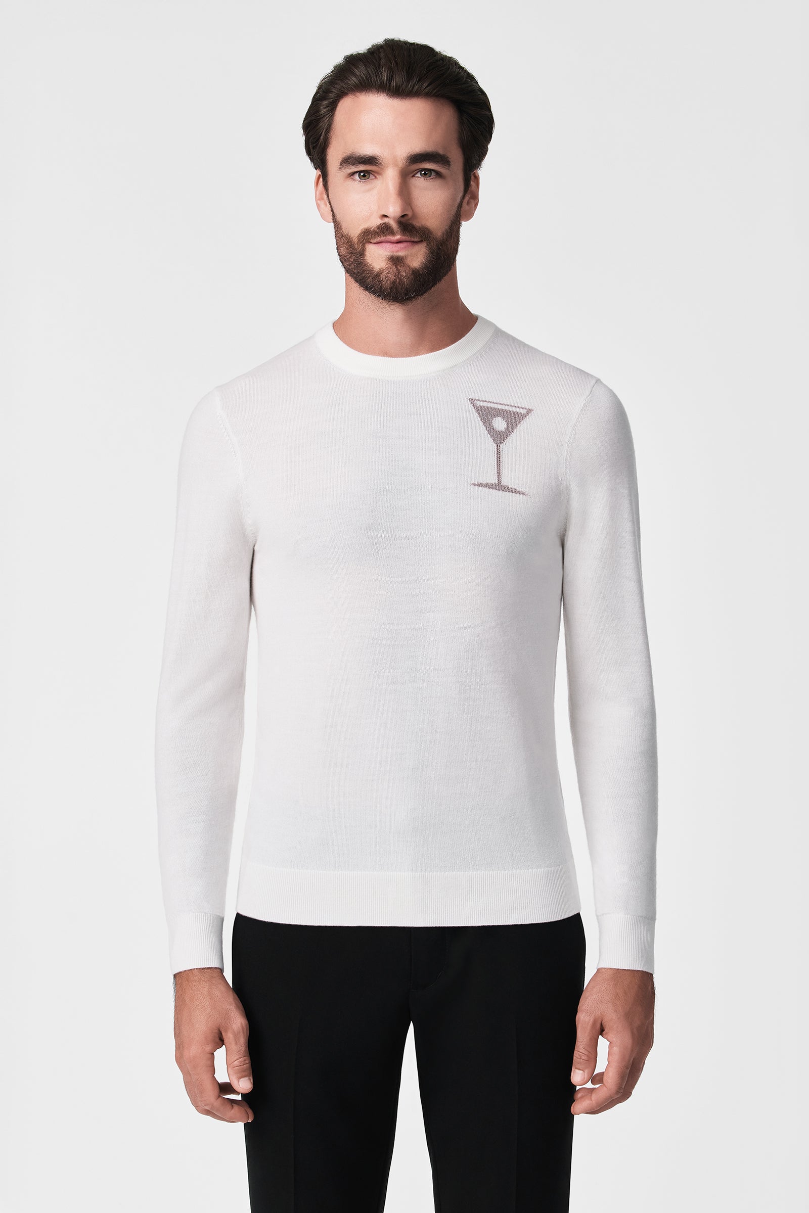 A man with dark hair and a beard wears the Martini Crewneck—a white long-sleeve sweater with a small martini glass graphic on the left chest—paired with black pants against a white background.