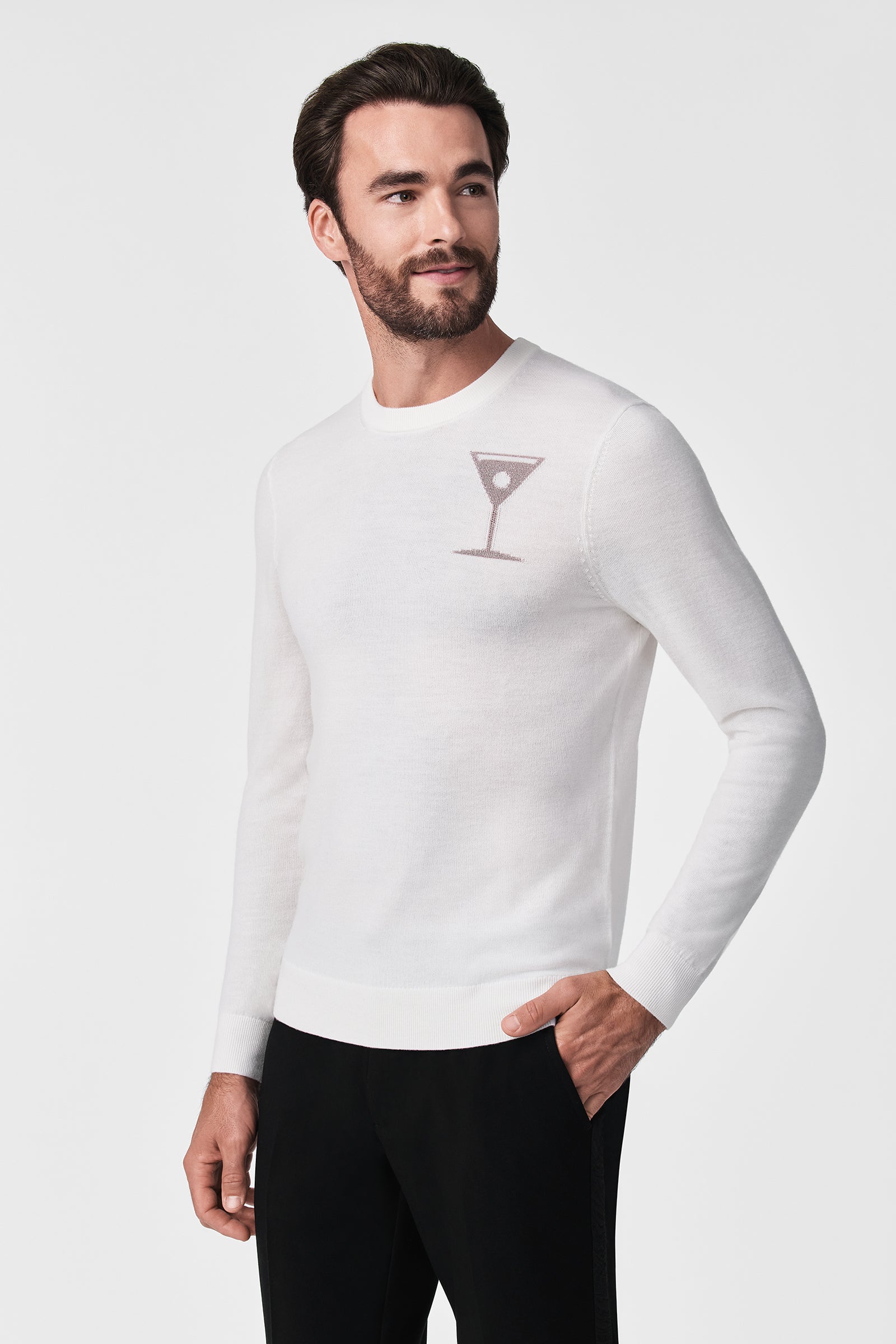 A man with short dark hair and a beard wears the Martini Crewneck, a white sweater with a small martini glass graphic on the chest, paired with black pants, standing against a plain white background.
