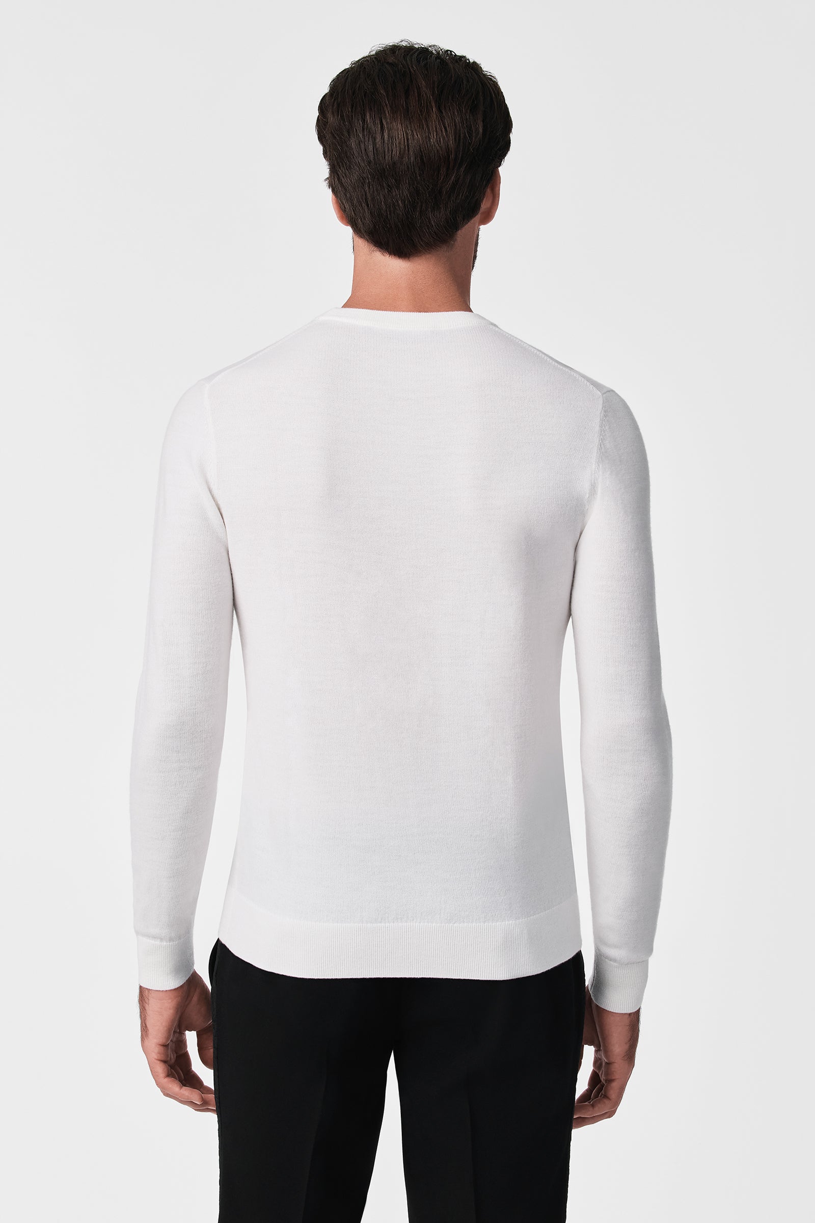 A man faces away from the camera, dressed in the Martini Crewneck long-sleeved white sweater and black pants, set against a plain white background.