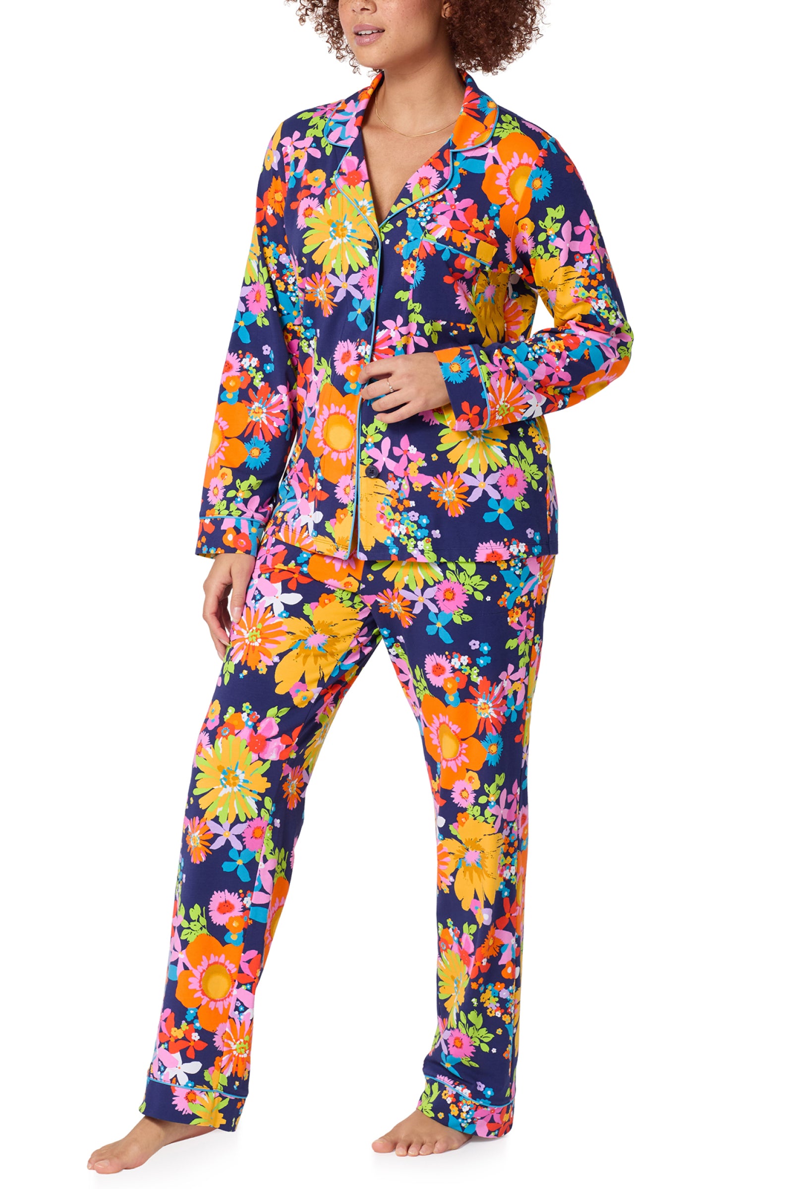 A person stands barefoot wearing the Classic PJ Set - Bay St Bloom, featuring a bold floral pattern in orange, yellow, pink, and green on a dark blue background. The pajama top has long sleeves and front buttons.