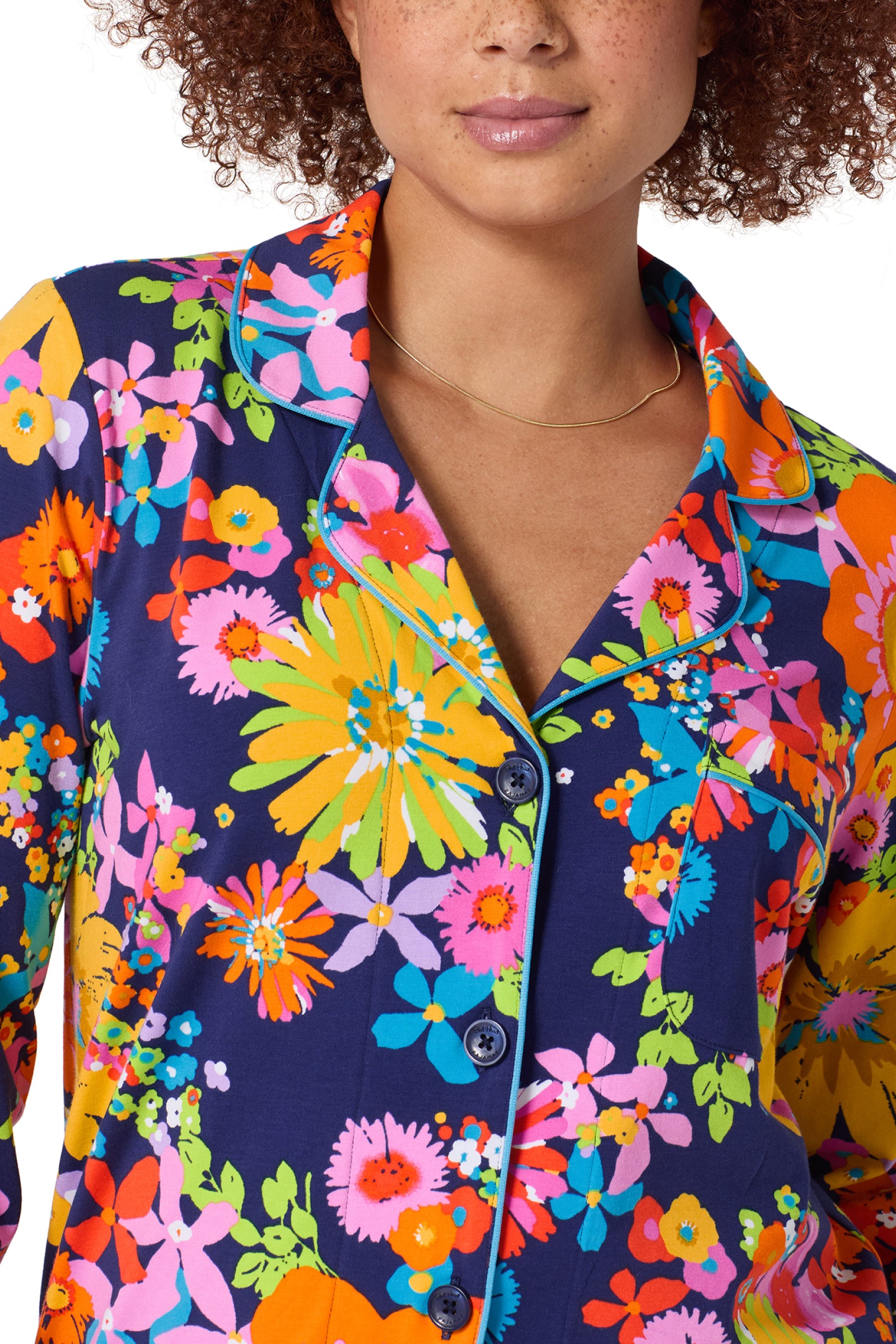 A person with curly hair wears the Classic PJ Set - Bay St Bloom: a navy pajama top with a vibrant floral pattern in yellow, orange, pink, and green, finished with teal piping on the collar and button placket.