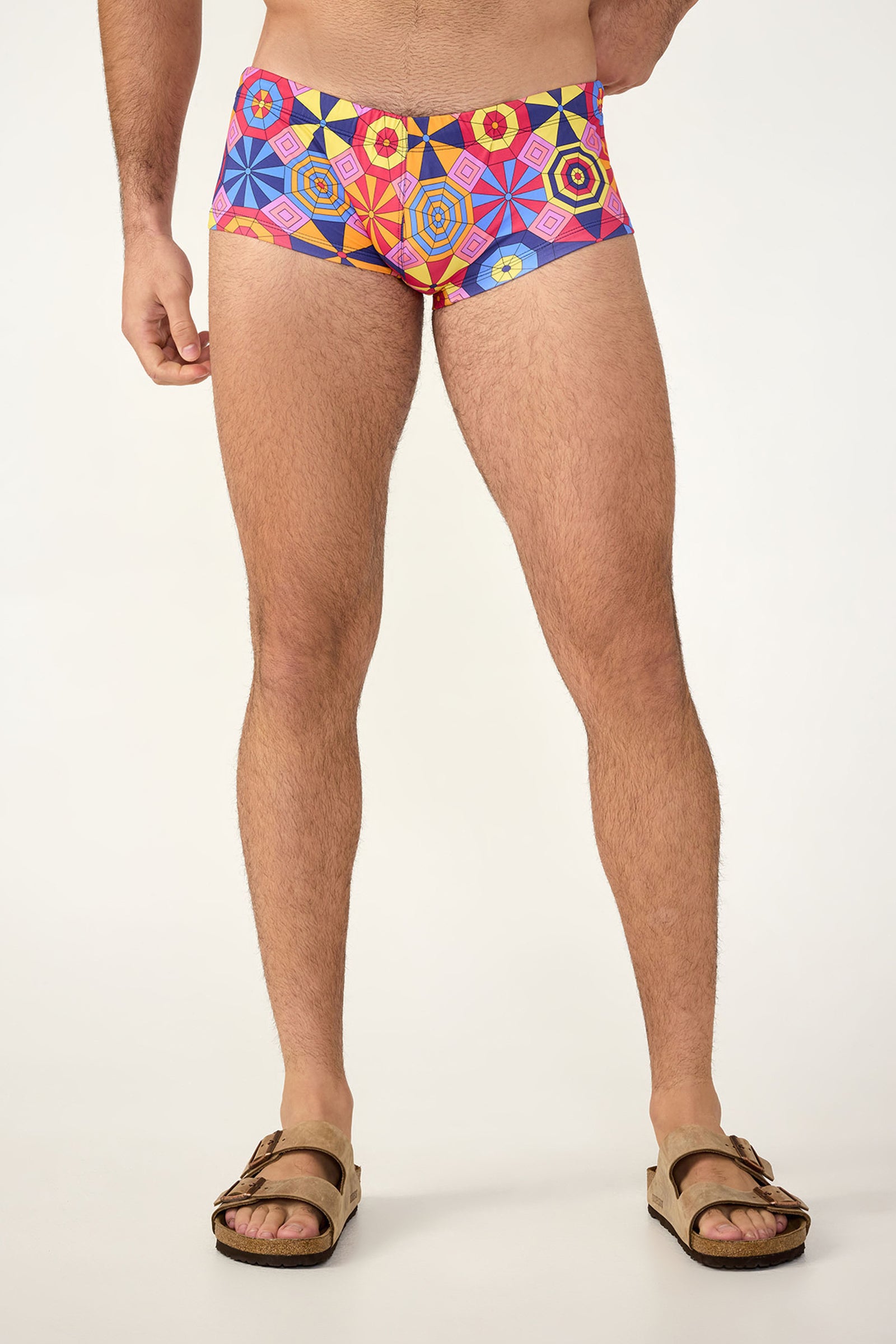 Only the lower torso and legs are visible as a person stands against a plain background wearing the South Beach Swim Trunk - Parasol in Multi, featuring colorful geometric patterns, paired with brown sandals.