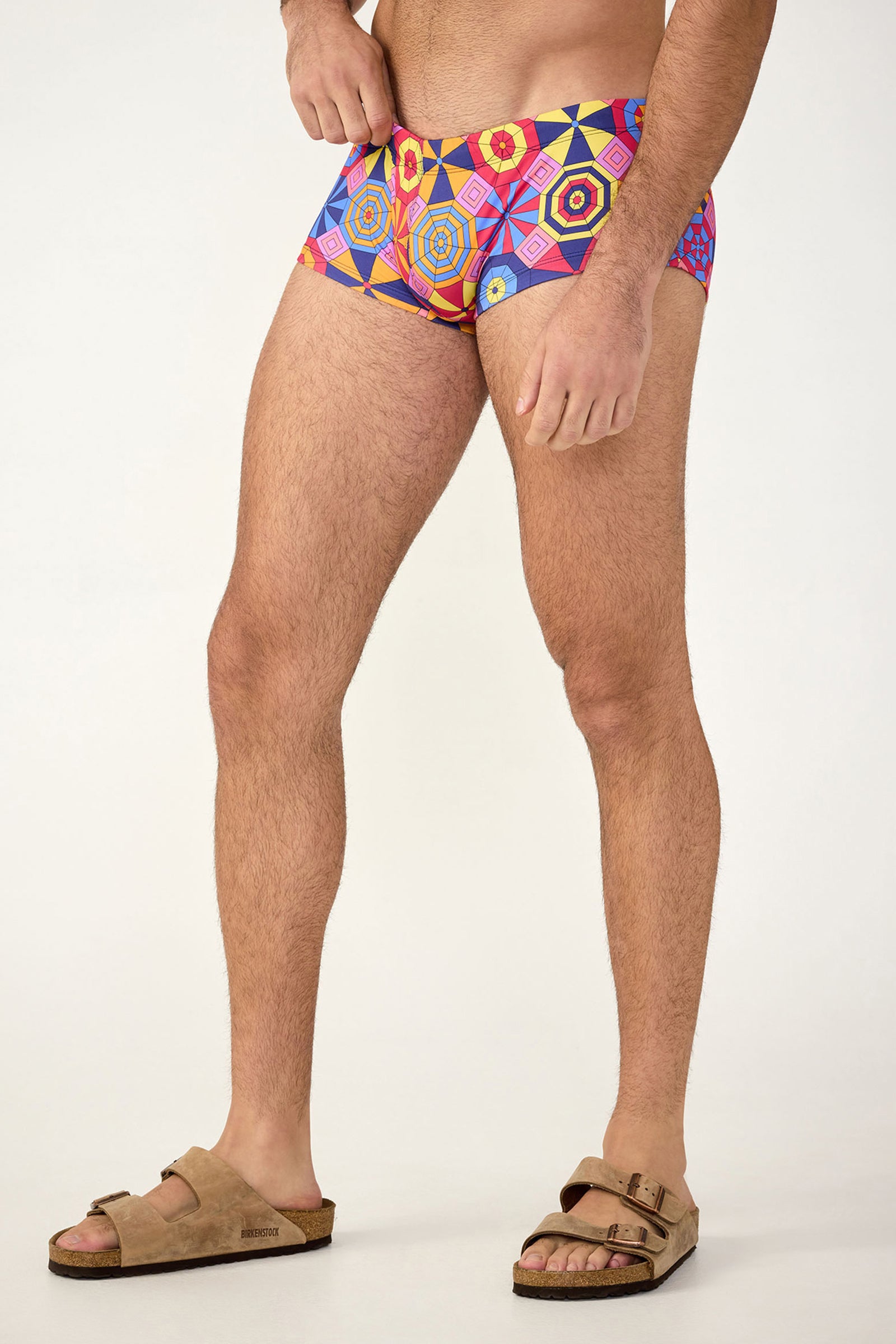 A man wearing South Beach Swim Trunk - Parasol, featuring colorful geometric patterns, and tan sandals stands against a plain light background; only his body from the chest down is visible.