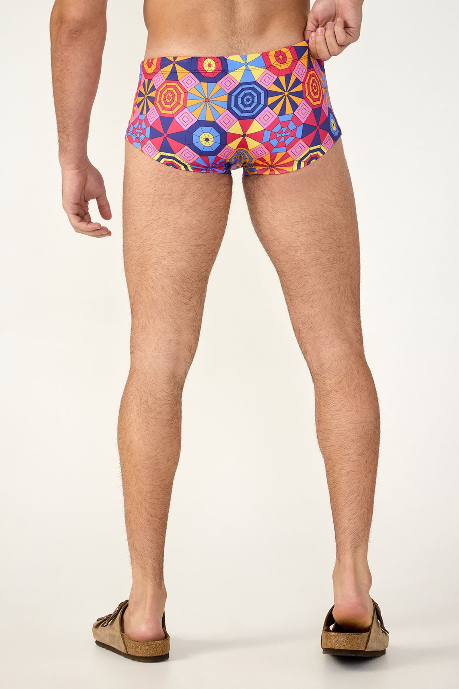 A person stands barefoot in brown sandals, seen from behind, wearing South Beach Swim Trunk - Parasol with a colorful geometric pattern of hexagons and bright shapes on a neutral background.